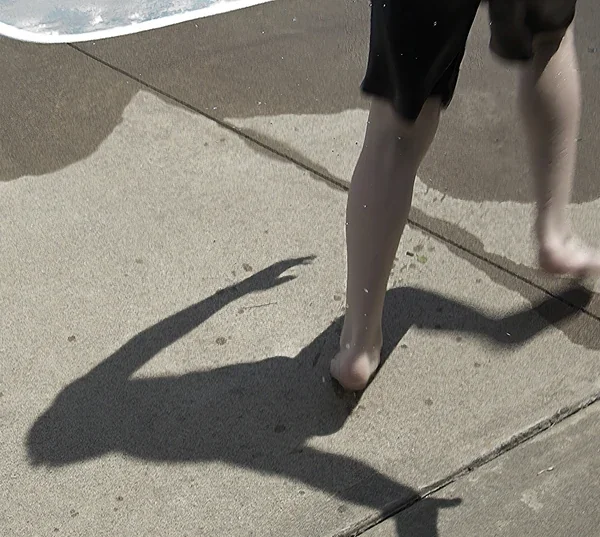 Todd walking barefoot on sunlit pavement near the water as his long shadow stretches dramatically across the ground.