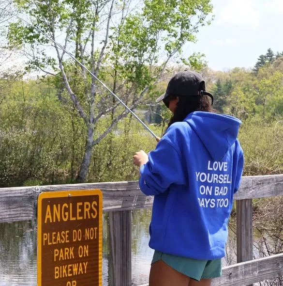 A person holds a fishing rod beside a railing, wearing a blue sweatshirt reading Love yourself on bad days too.