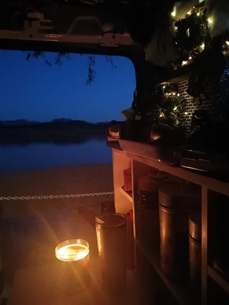 Candle and Christmas lights glow inside a small camper van at dusk, with the Colorado River visible through the open doorway.