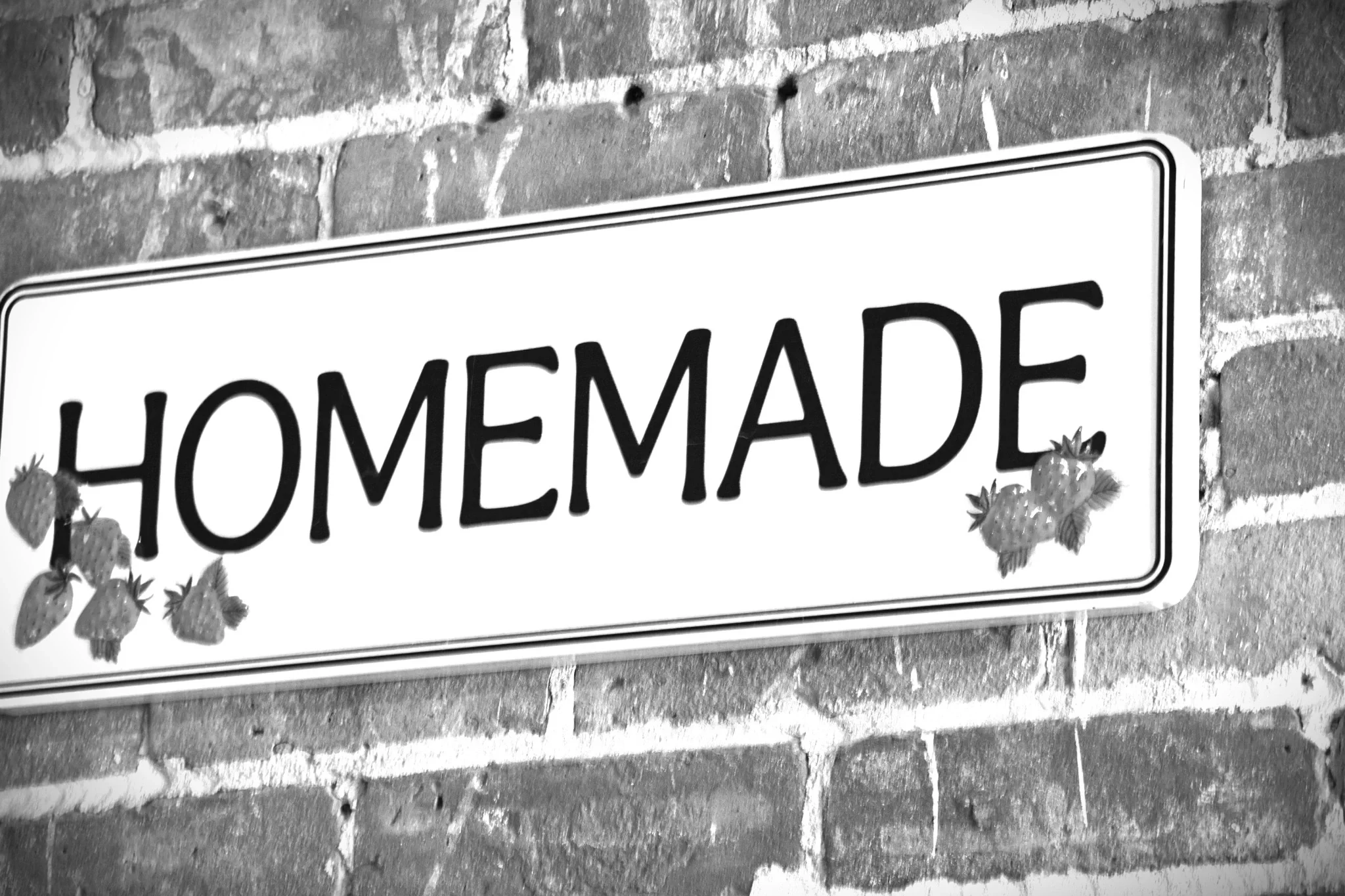 Black-and-white photograph of a professionally manufactured HOMEMADE sign decorated with strawberries and mounted on a brick wall.