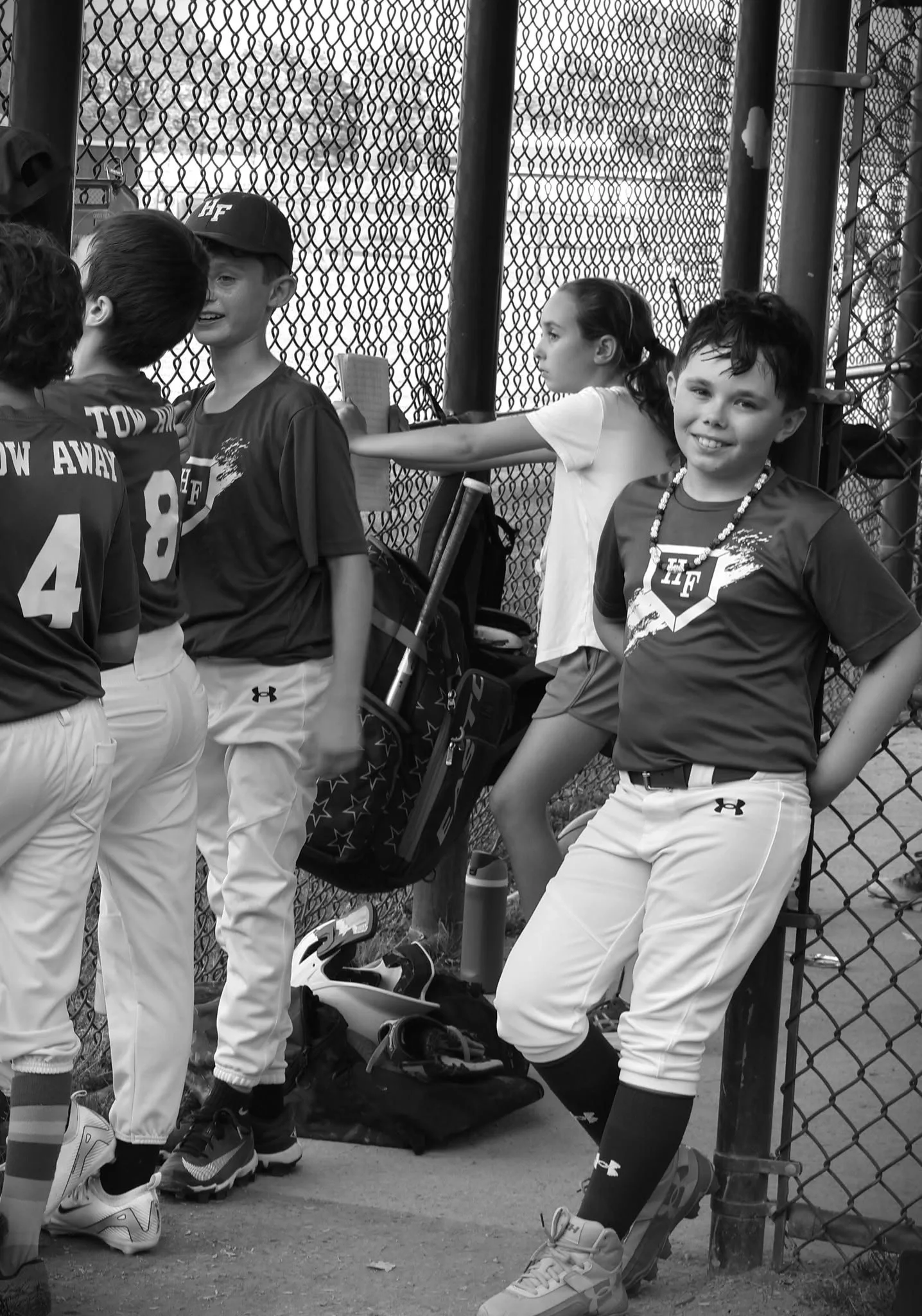 Black-and-white photograph of a young baseball player smiling toward the camera while teammates gather casually inside a chain-link dugout.