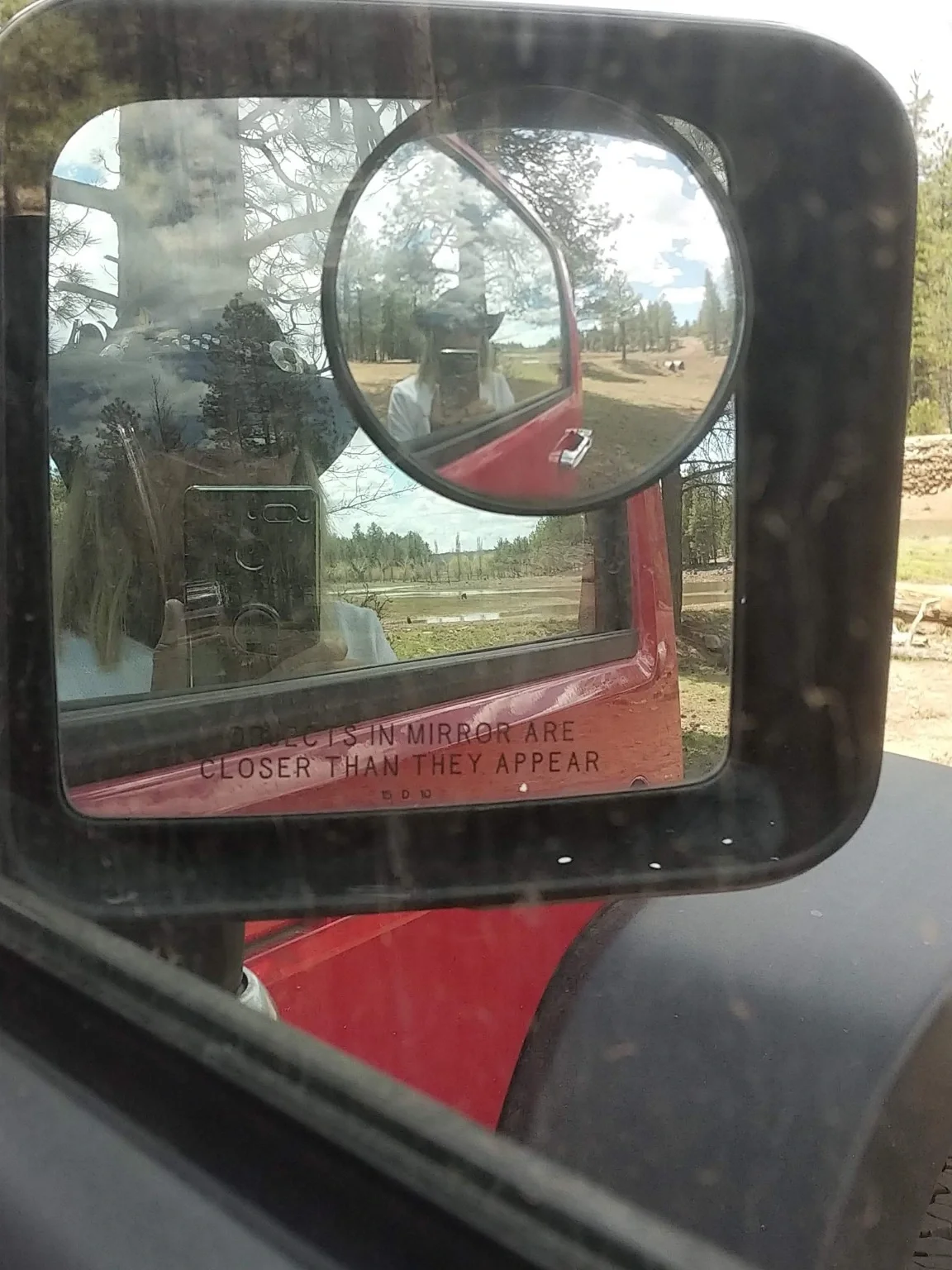 Photographer reflected in the side mirrors of a red vehicle with a wooded landscape behind her.