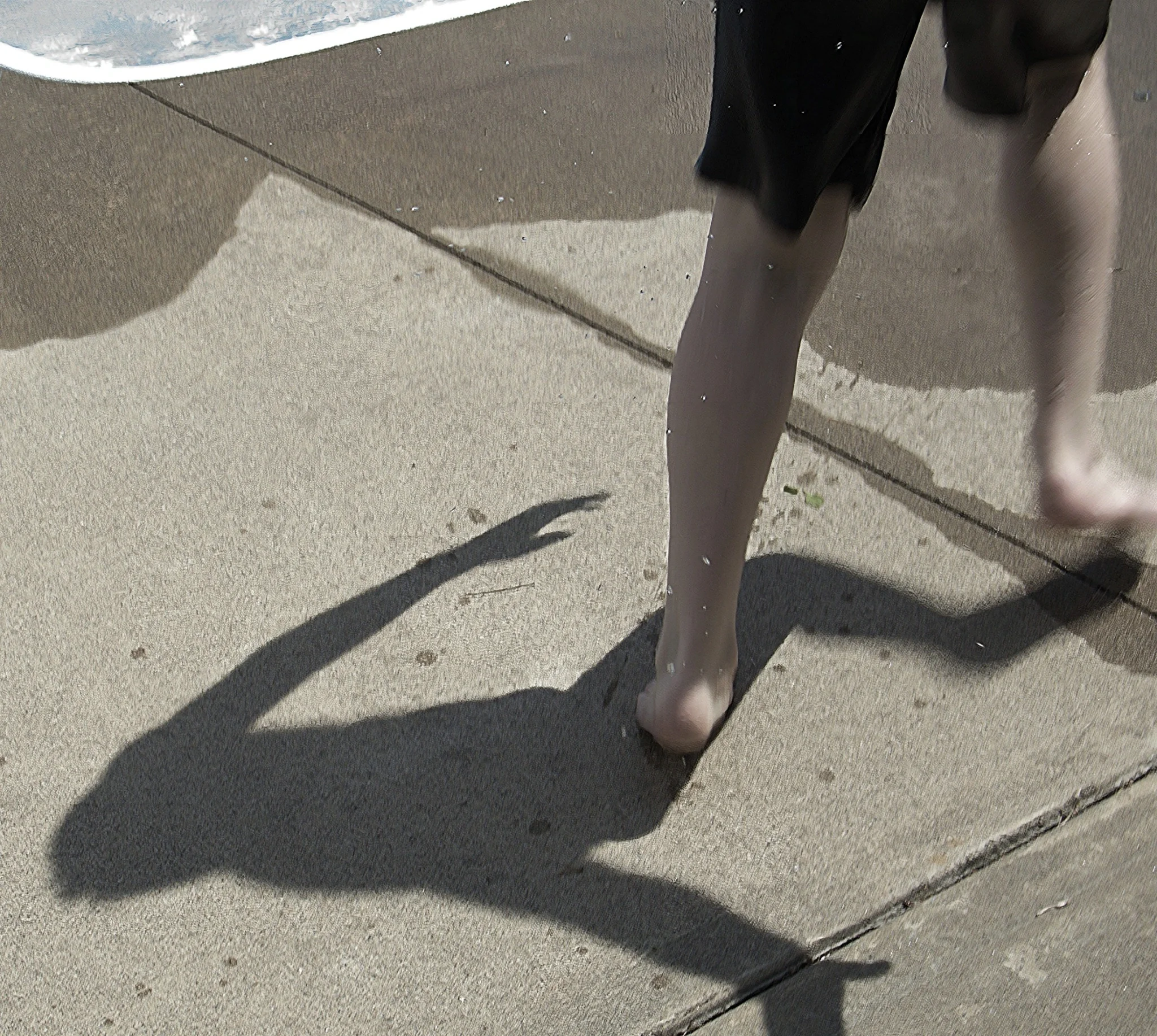 Todd walking barefoot on sunlit pavement near the water as his long shadow stretches dramatically across the ground.
