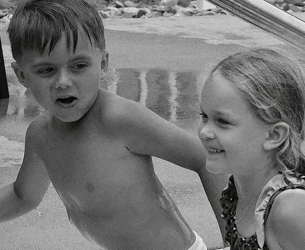 Black-and-white candid photograph of a young boy in the water looking toward a smiling young girl beside him.