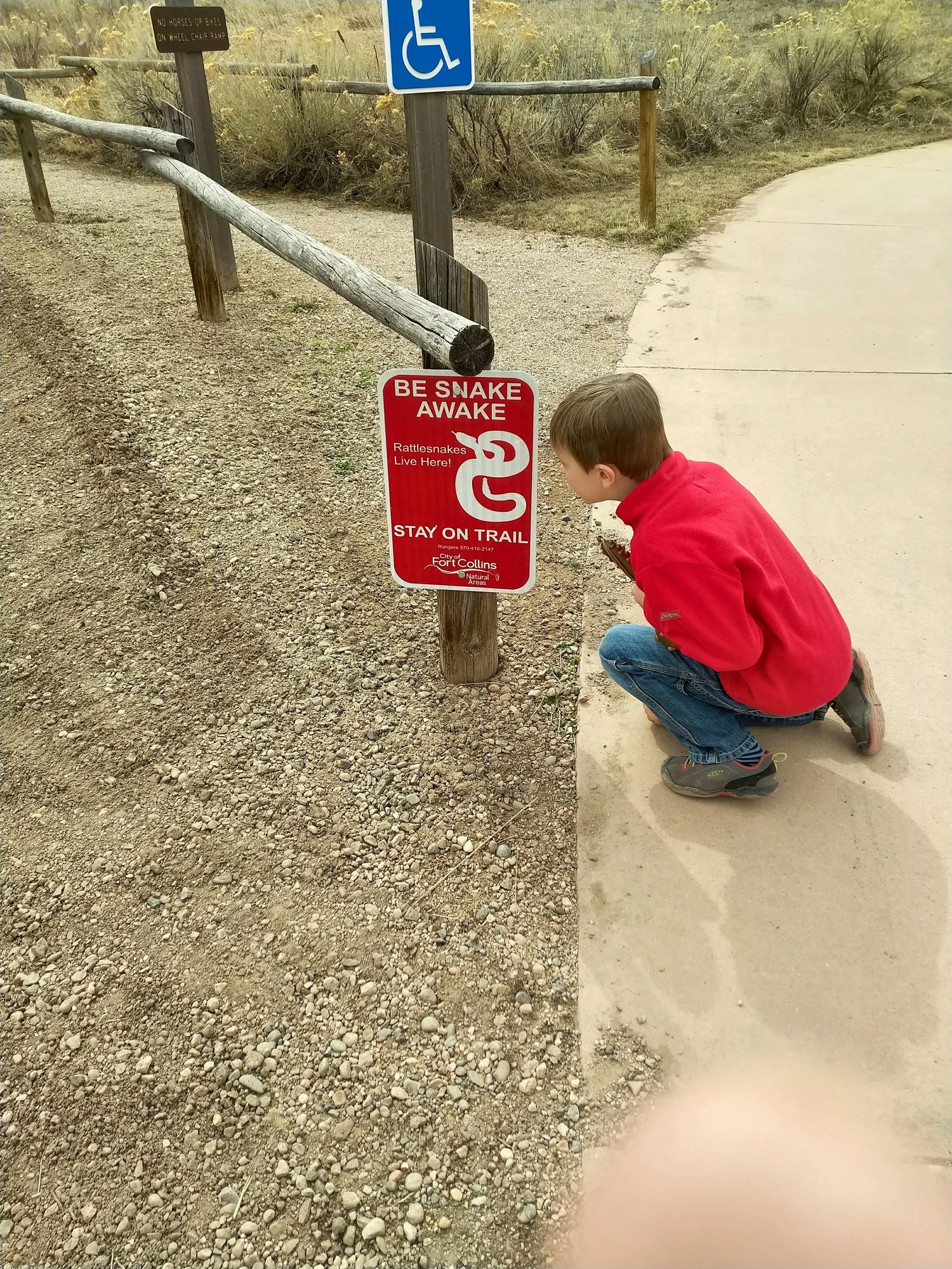 Child crouching beside a red Be Snake Awake sign warning that rattlesnakes live here and to stay on the trail, along a paved trail in Colorado.