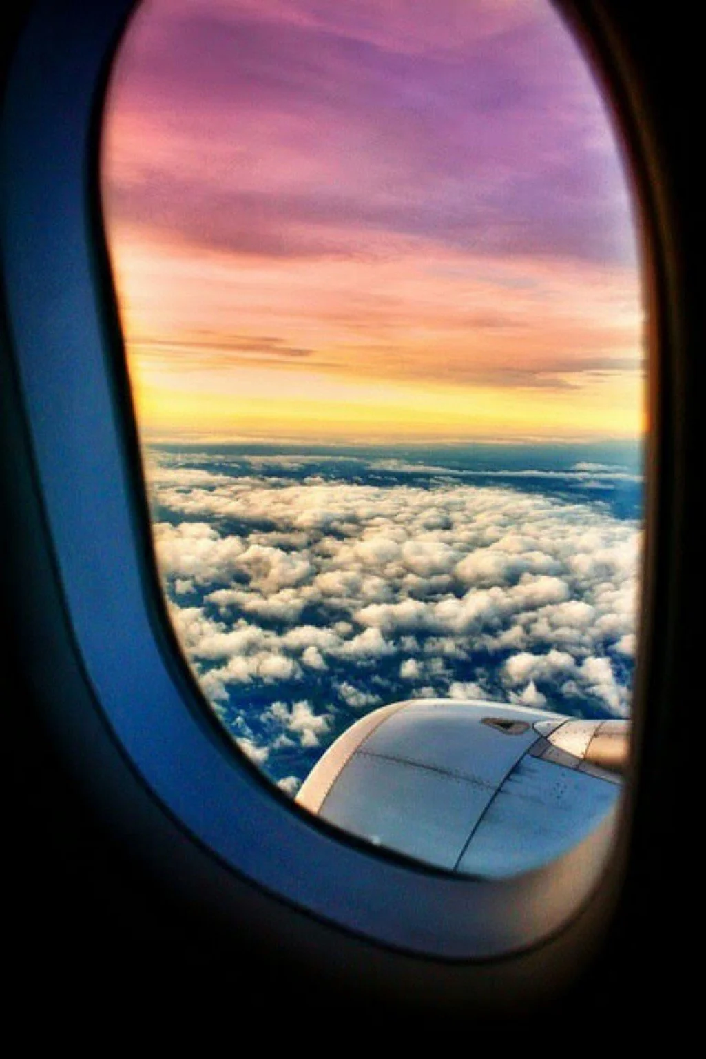 View through an airplane window of a pink, purple, and gold sky above a wide blanket of clouds, with the aircraft engine visible below.