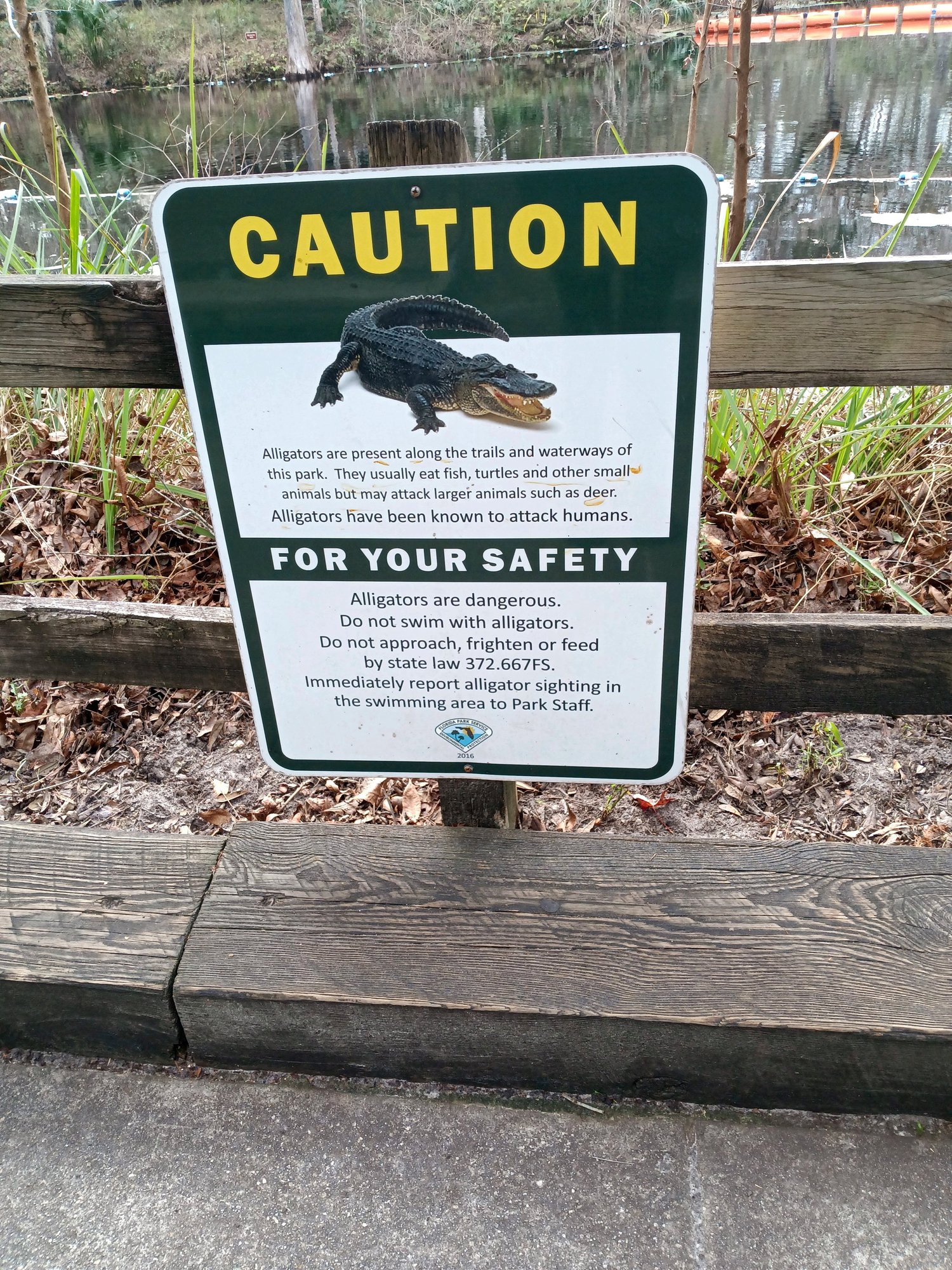 Florida park caution sign warning that alligators are present along the trails and waterways, posted on a wooden fence beside the water.