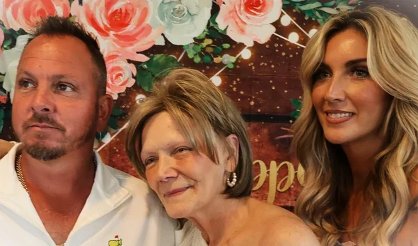 Three adults pose together in front of a floral birthday backdrop.