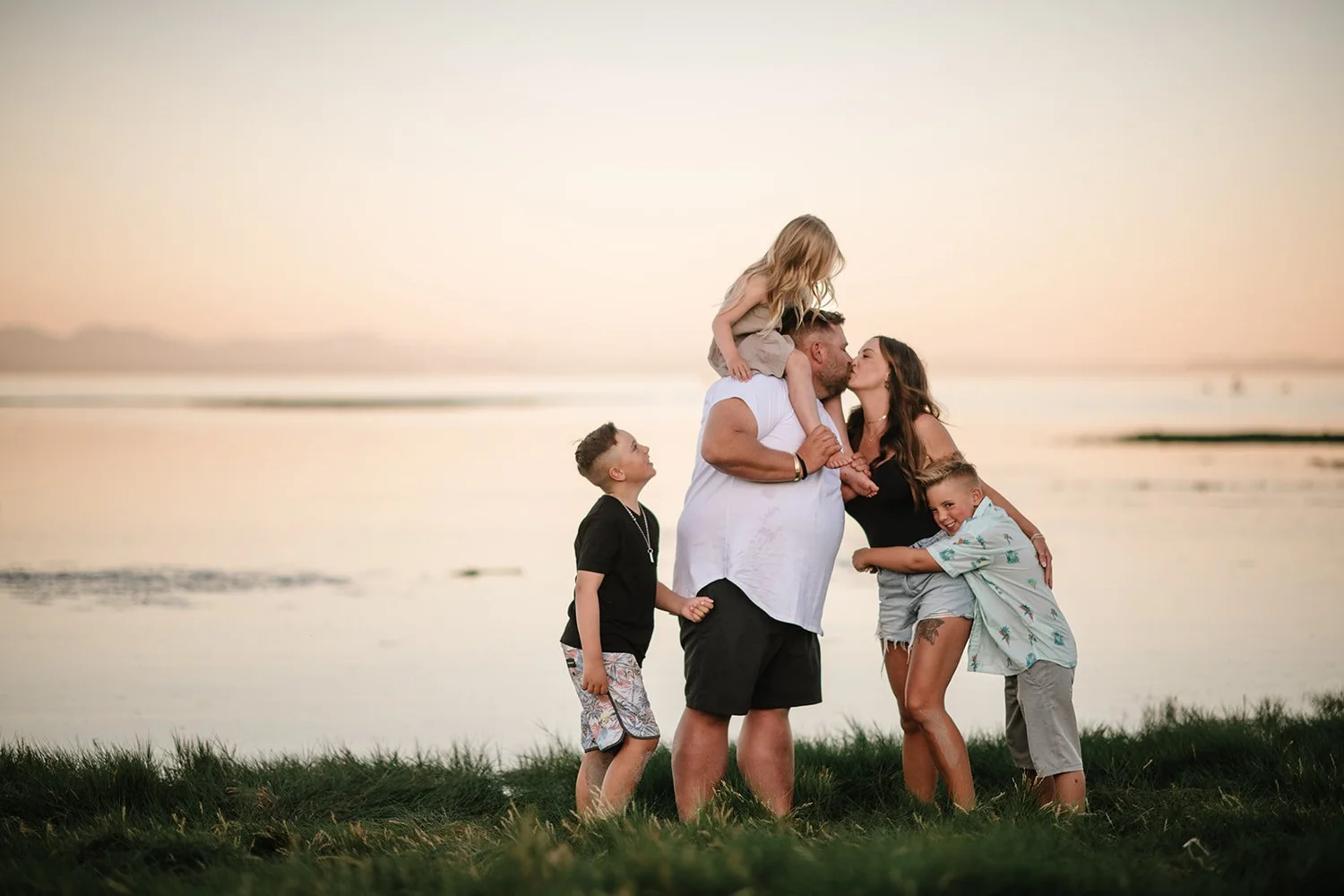 Parents sharing a kiss by the ocean at dusk, their daughter on dad's shoulders and their two boys hugging close