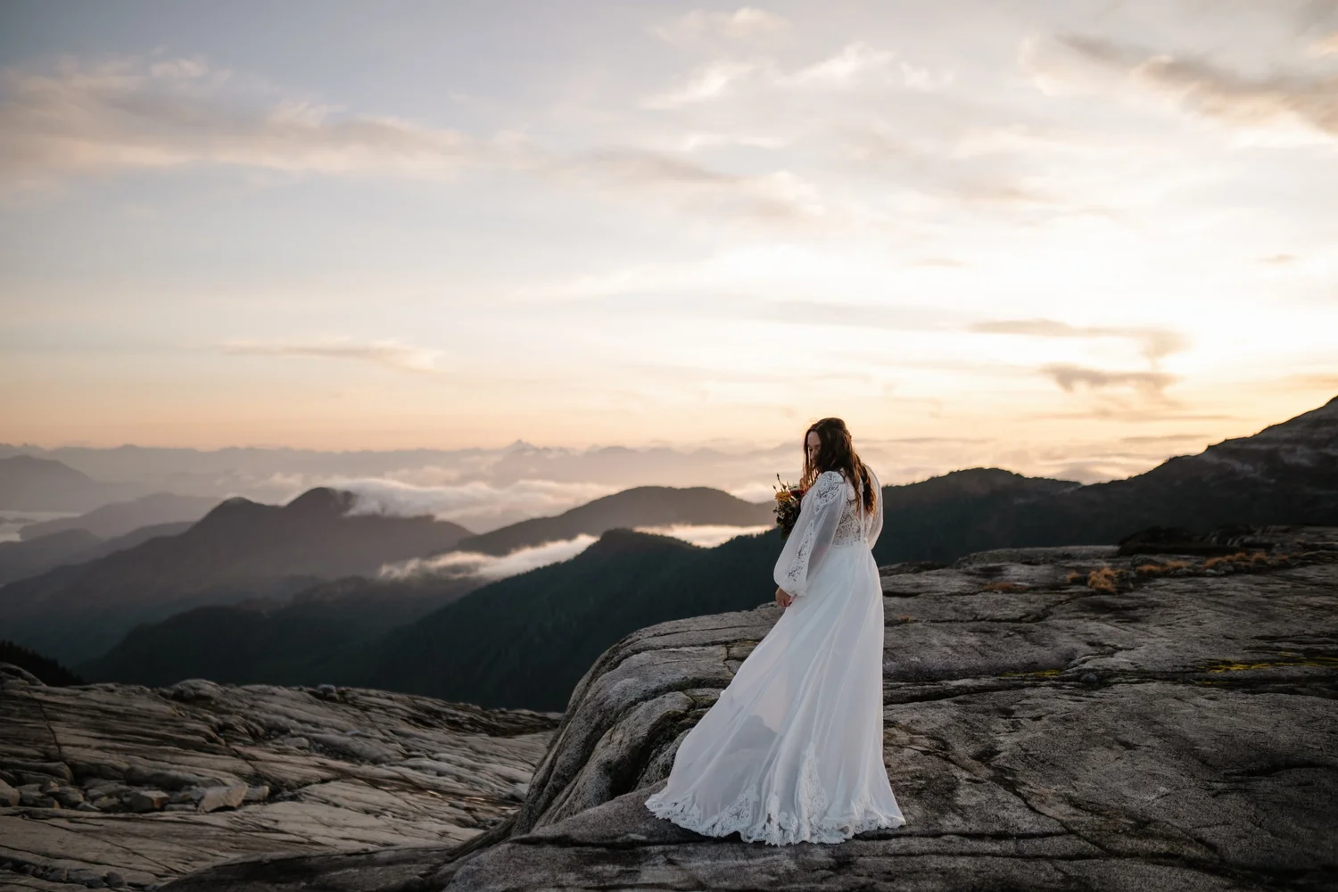 A bride standing alone above a sea of clouds at sunset