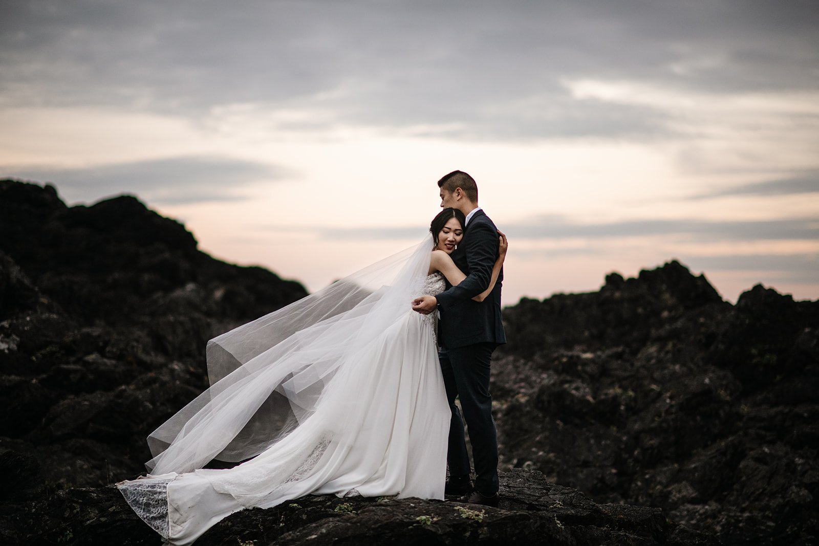 A bride and groom on dark rocks under a moody sky as her veil streams in the wind
