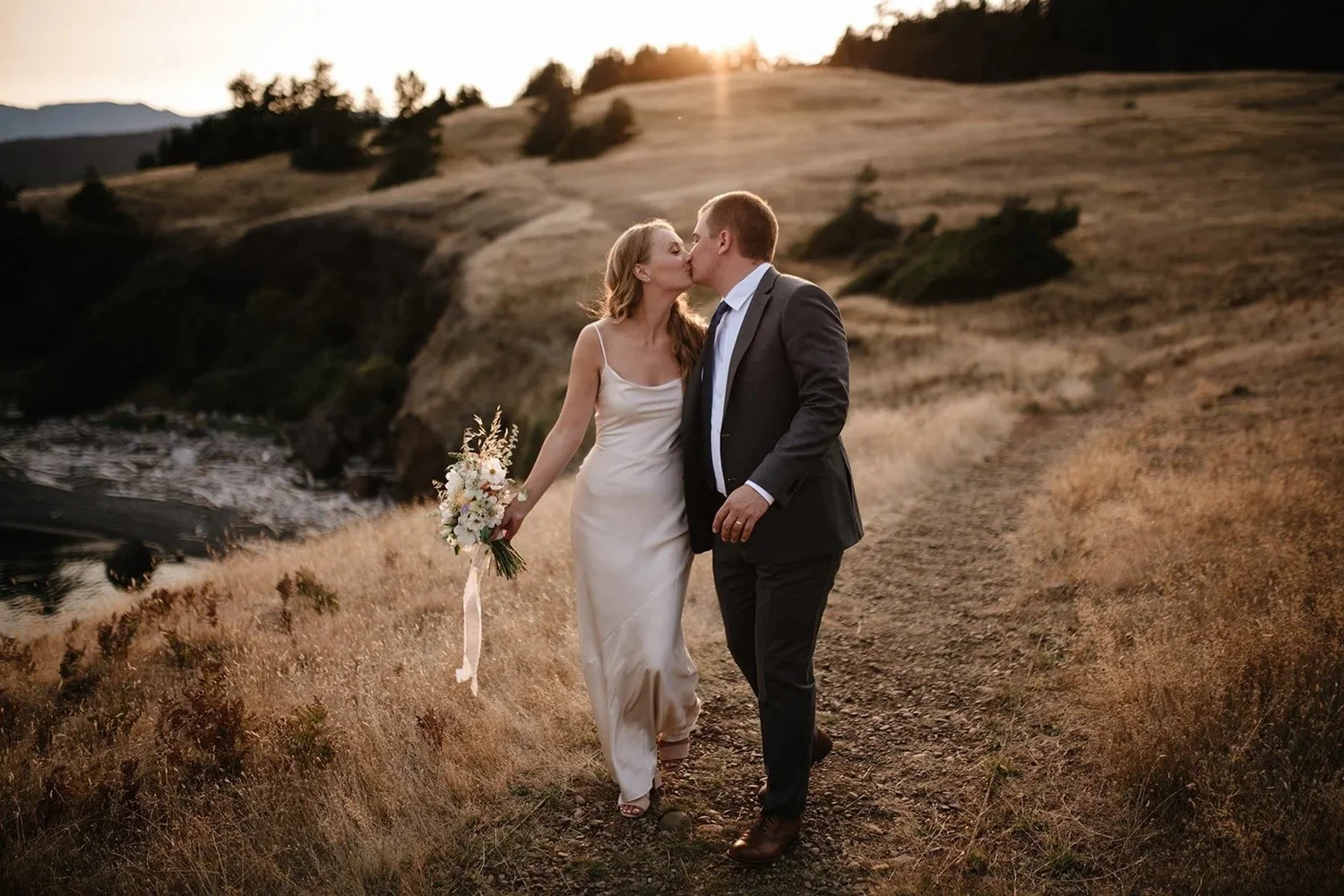 A bride and groom kissing in golden grass at sunset