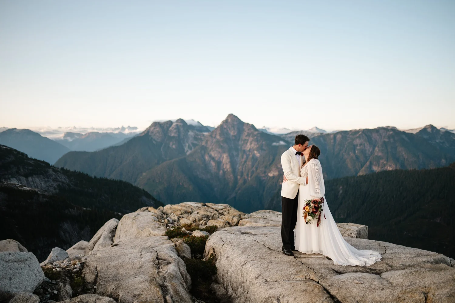 A couple kissing on a rocky outcrop above a mountain valley