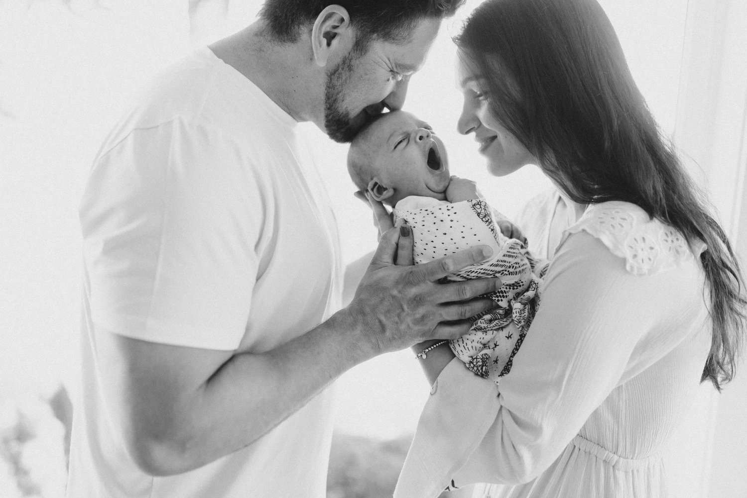 New parents holding their yawning newborn close, in black and white