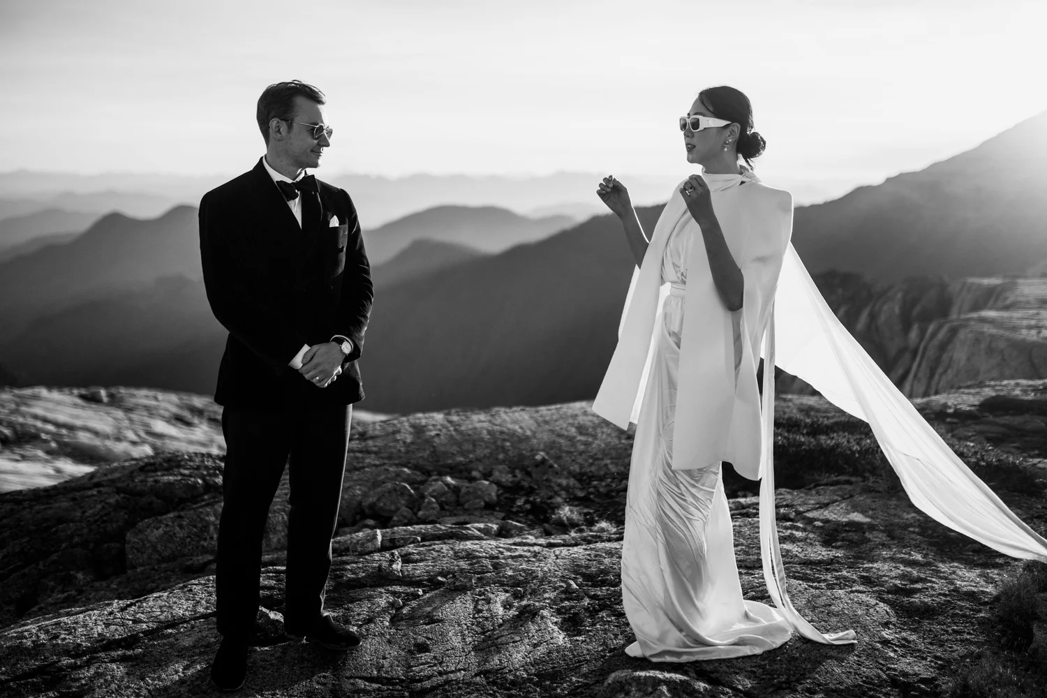 A groom and bride on a mountaintop, her long veil lifting in the wind, in black and white