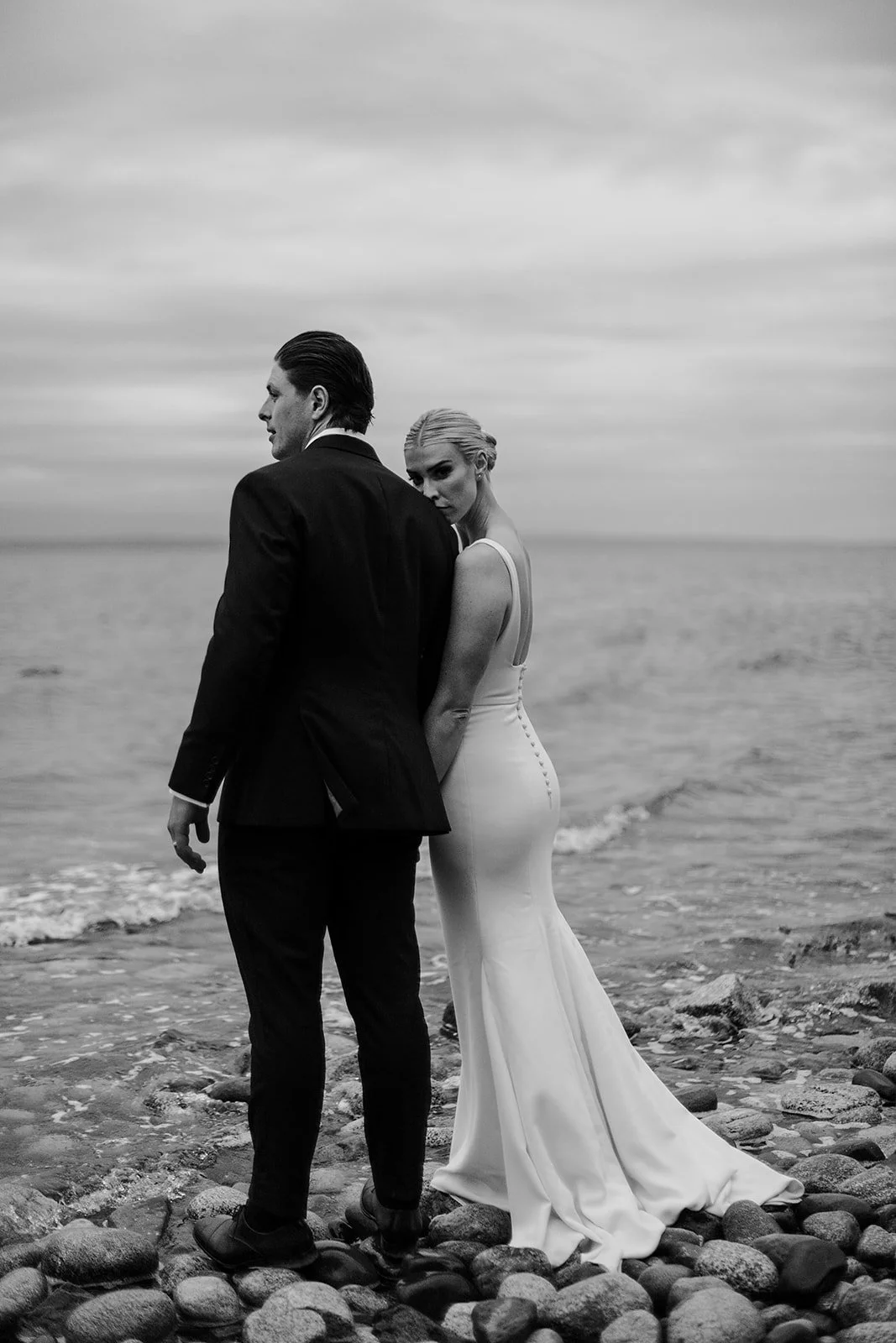 A bride and groom standing together on a pebble beach