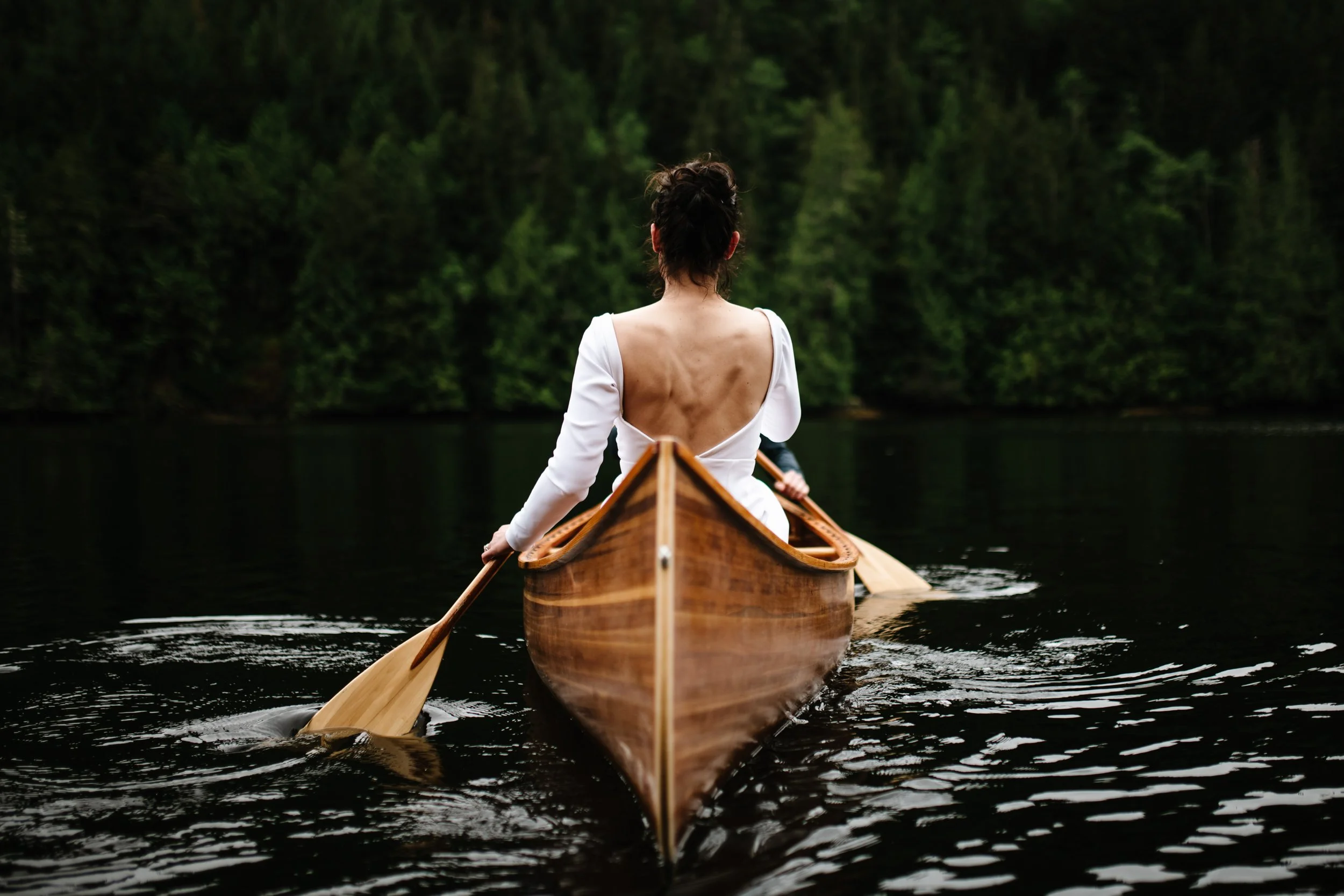A bride paddling a cedar canoe toward a dark wall of forest on Vancouver Island