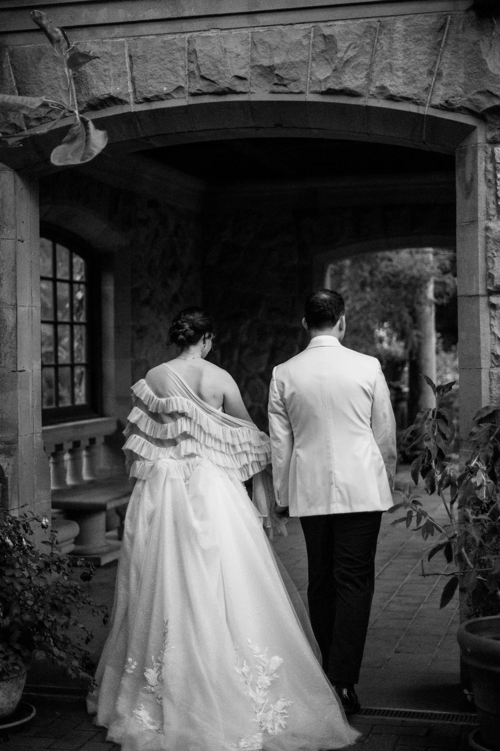 A bride and groom walking away together beneath a stone archway, in black and white