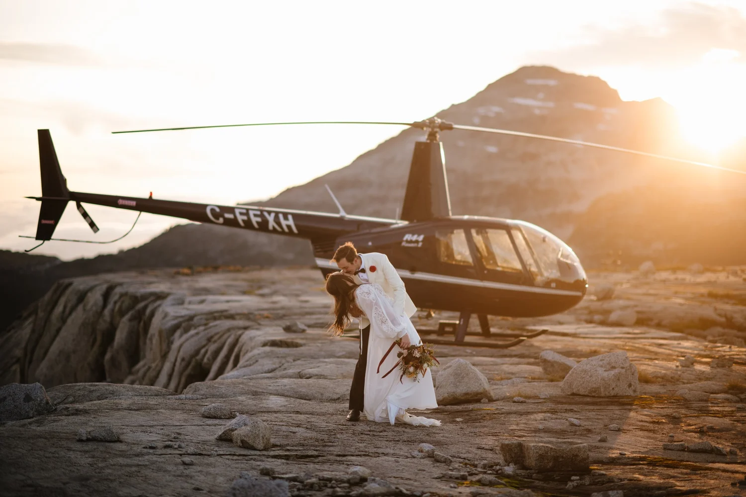 A couple kissing beside a helicopter on a mountaintop at sunset