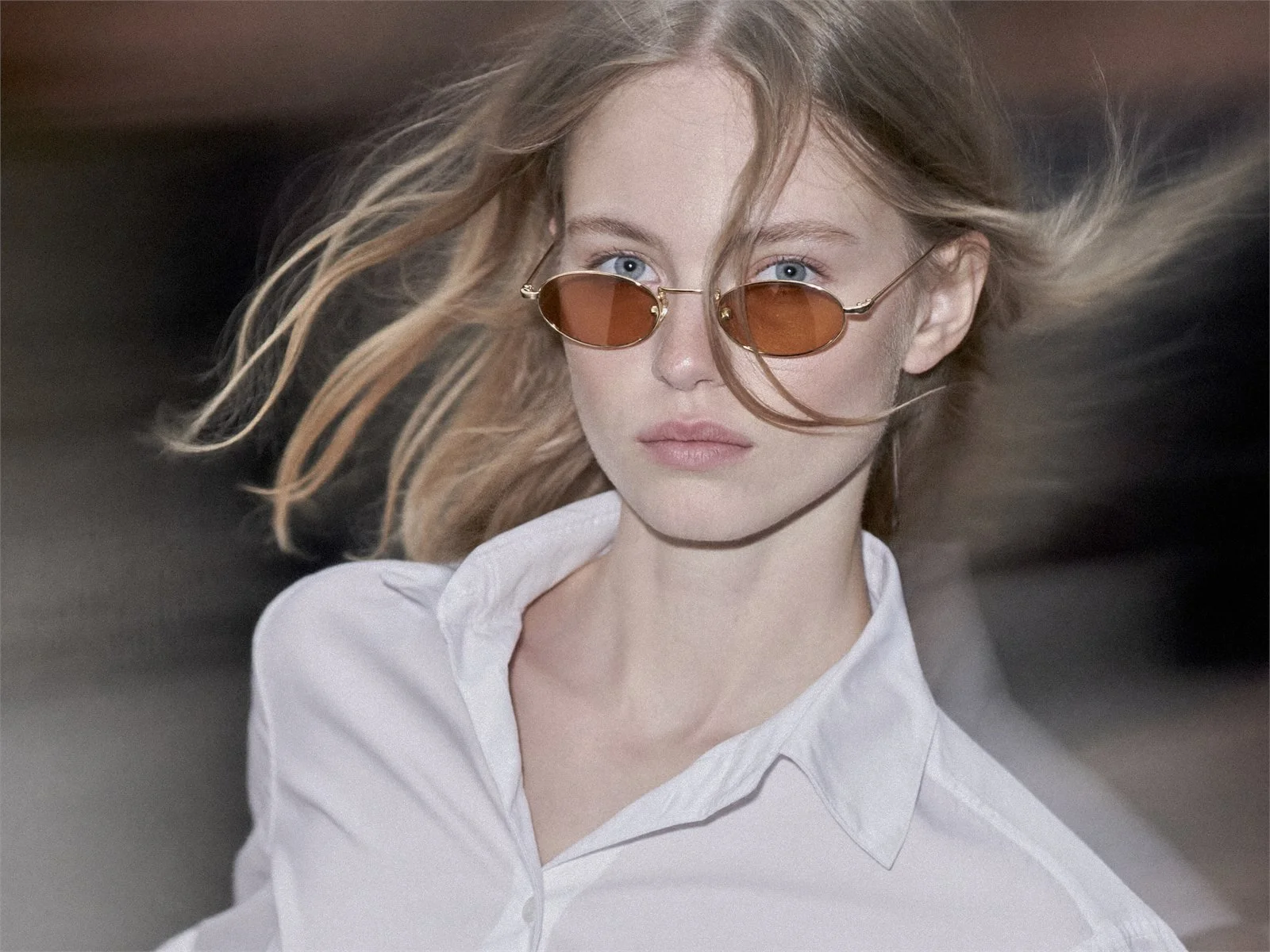 A woman with windblown hair in round vintage sunglasses.
