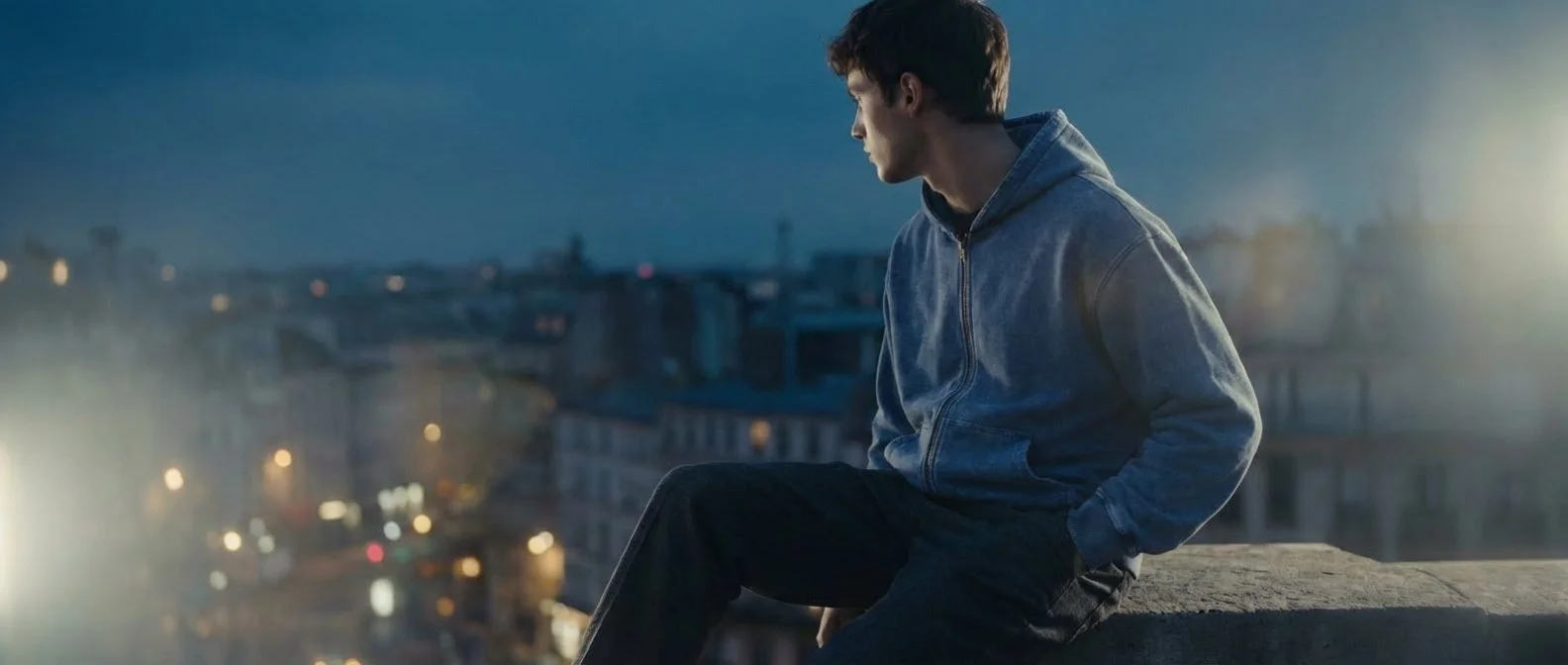 A figure seated on a rooftop ledge at night, the city lights blurred out behind him.