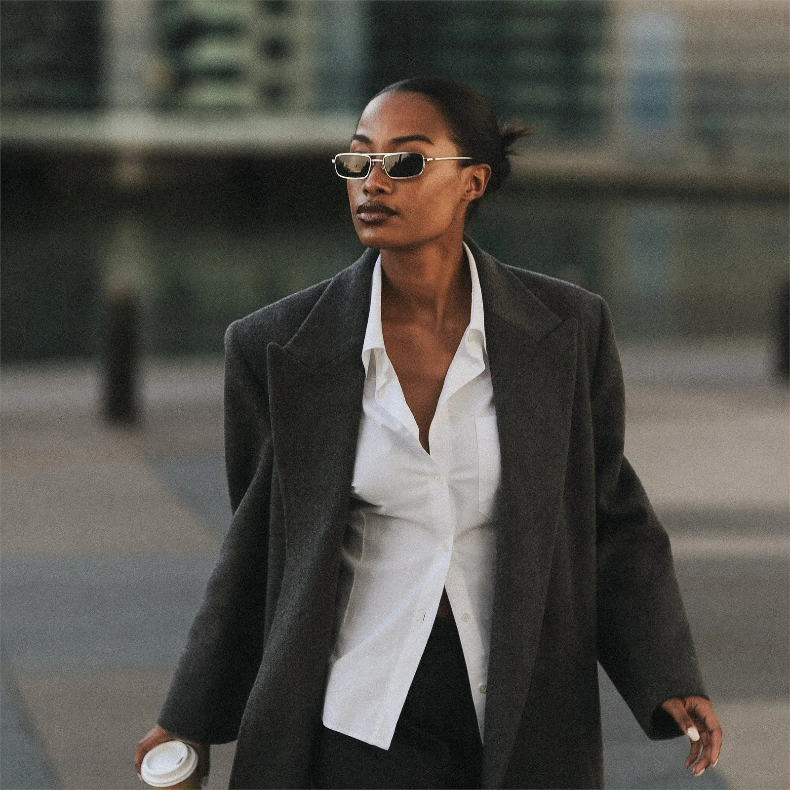 A woman in a dark wool coat and narrow sunglasses crossing a city street with a paper cup.