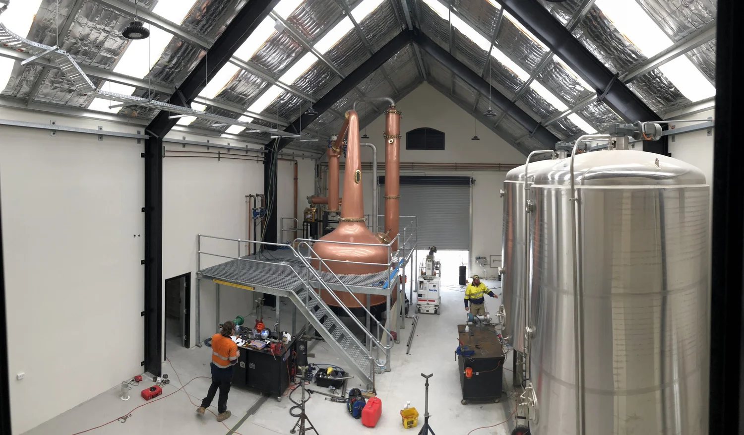The still house under a tall gabled roof with skylights: copper pot and column stills on a steel platform, stainless steel tanks alongside