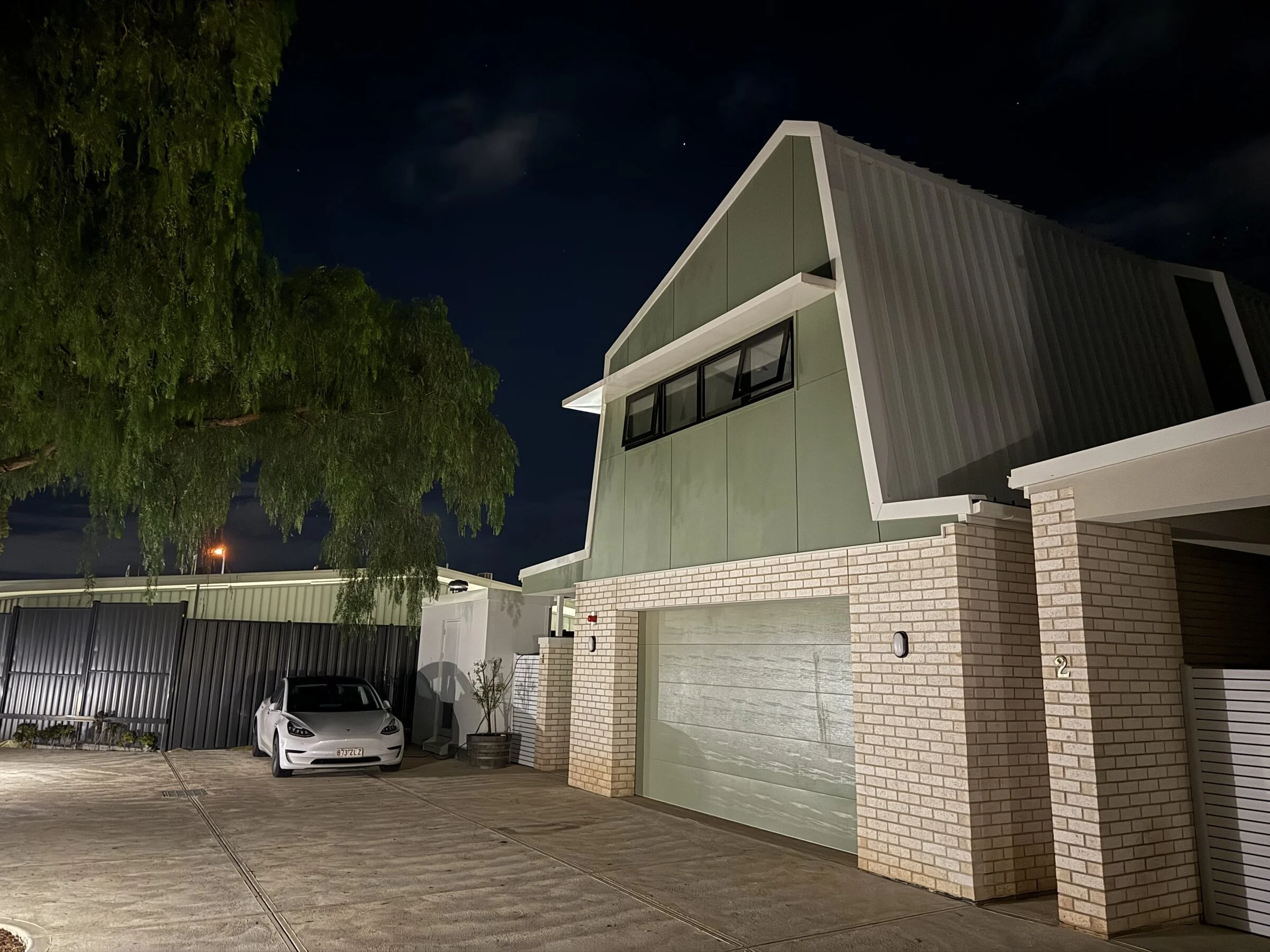 A townhouse at night: pale green cladding over a cream brick base, folded roof and clerestory windows, lit along the shared driveway