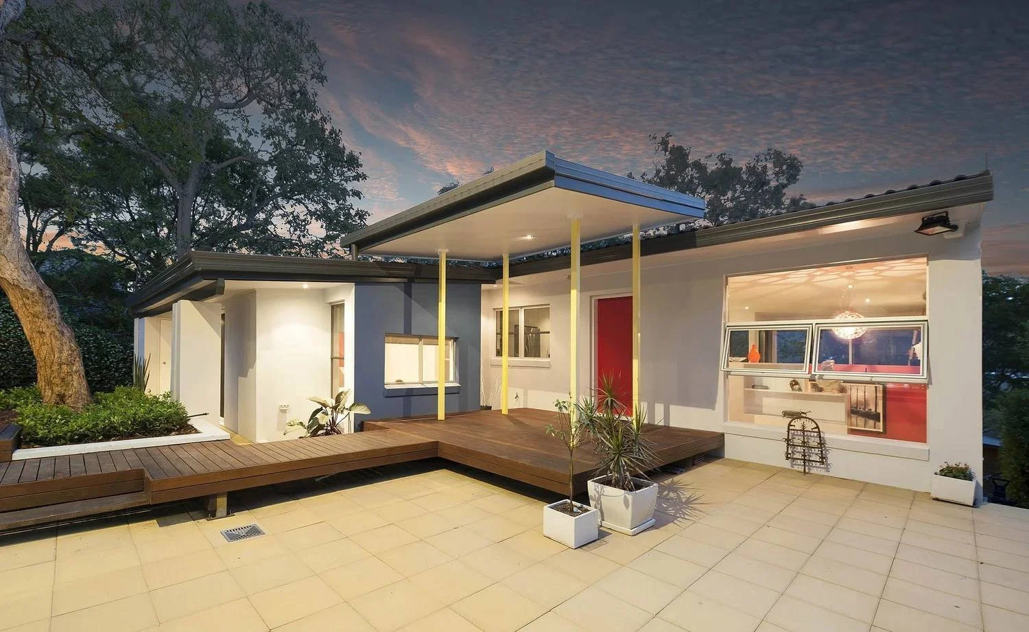 The renovated house at dusk, with a timber deck, a floating entry canopy on yellow posts and a red front door