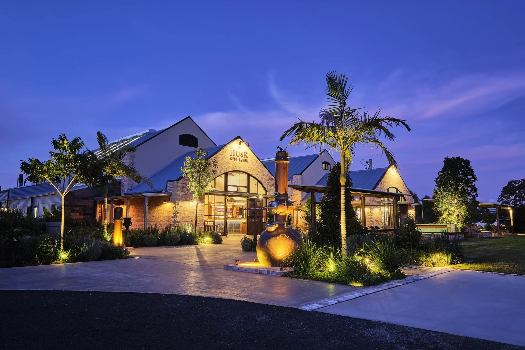 Husk Distillers at dusk: brick and white gables, an arched entry and a copper still among palms