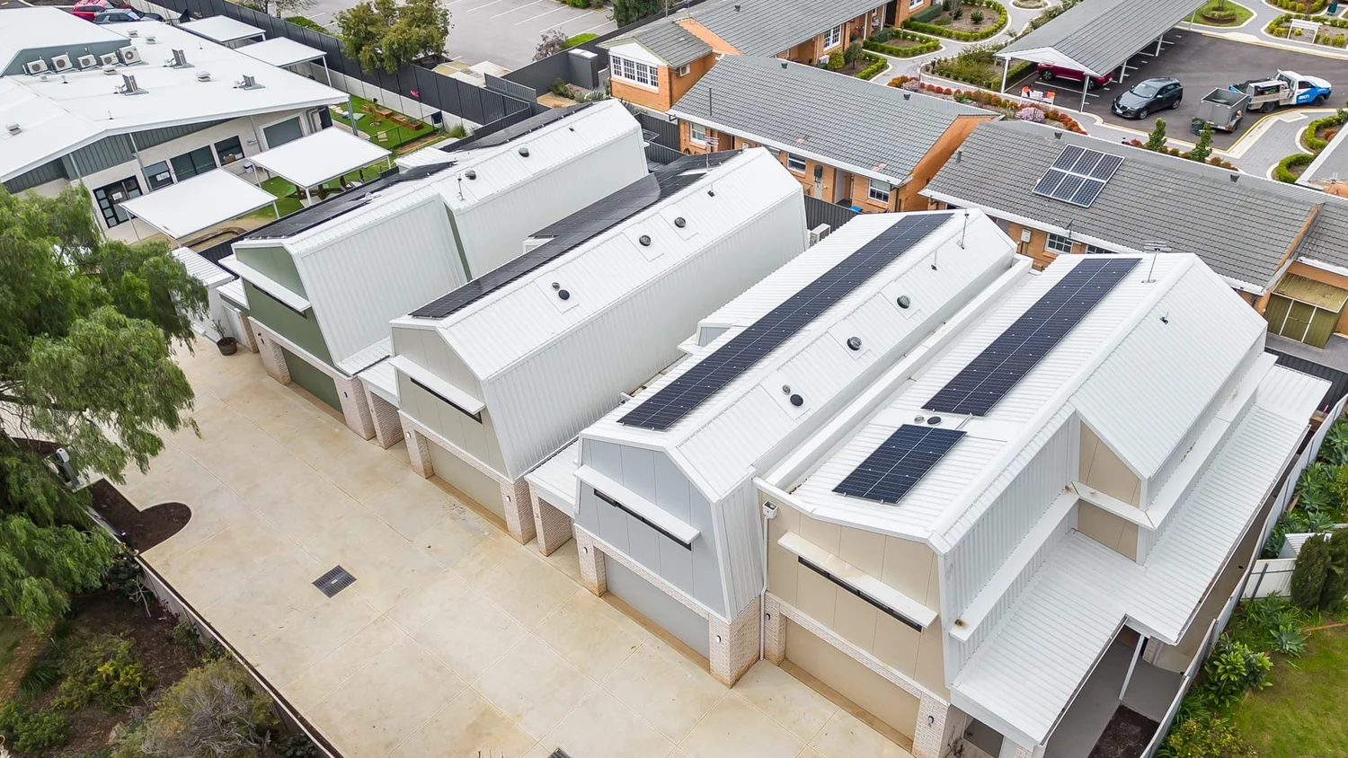 The four townhouses from above the driveway: folded roofs with solar panels over facades in green, white, grey and cream, with garages below