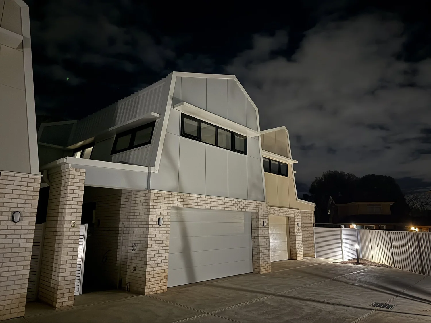Two townhouses at night with clerestory windows under folded roofs, the garages and entries opening off a level driveway