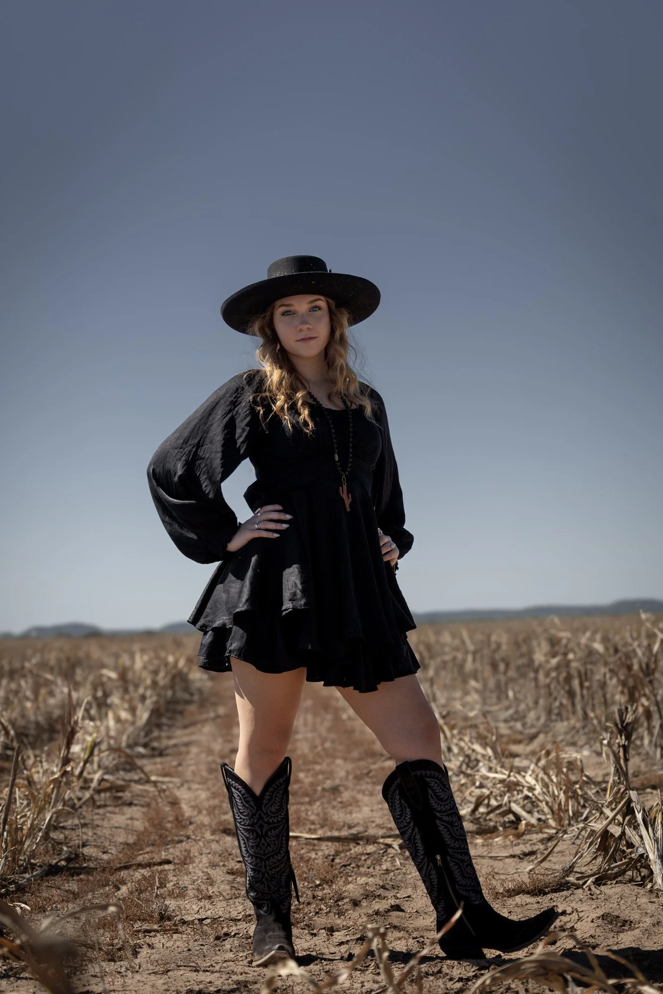 A person in a black dress, wide-brimmed hat, and patterned boots standing with hands on hips in a harvested field.
