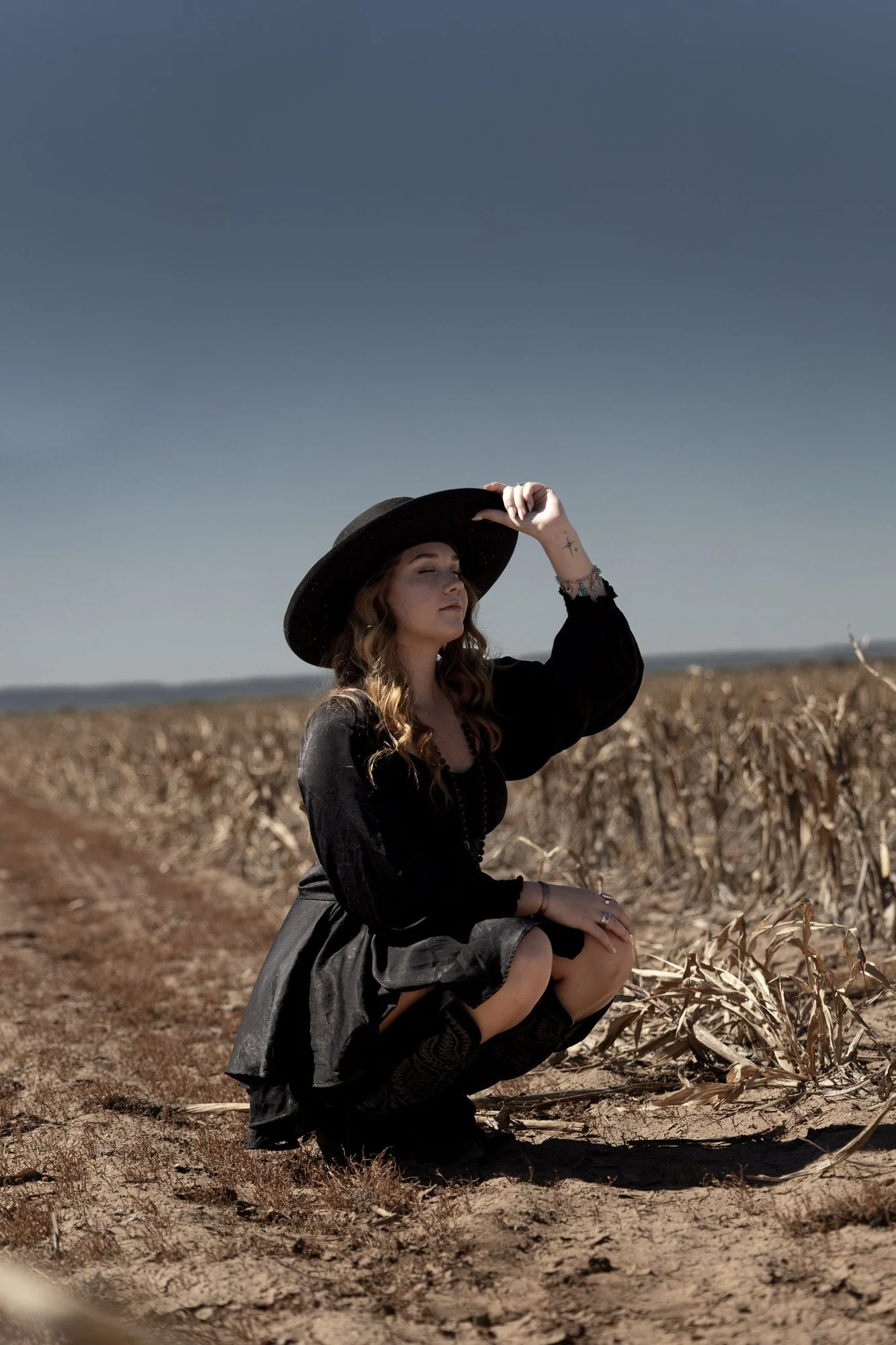A person crouching in a harvested field in a black dress and boots, with one hand on a wide-brimmed hat.