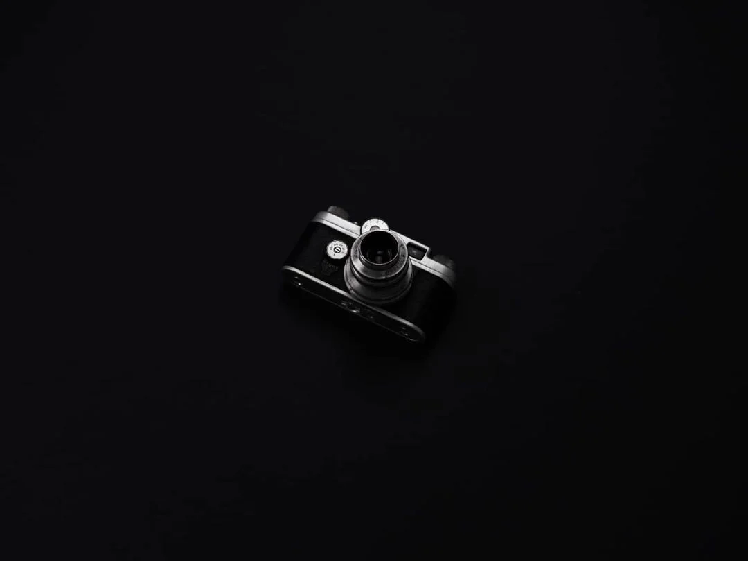 A small vintage camera photographed in black and white against a dark background.