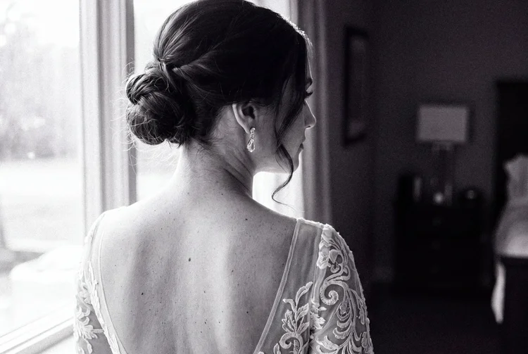 A bride with her hair in a low bun looks toward a window, lace across her open back