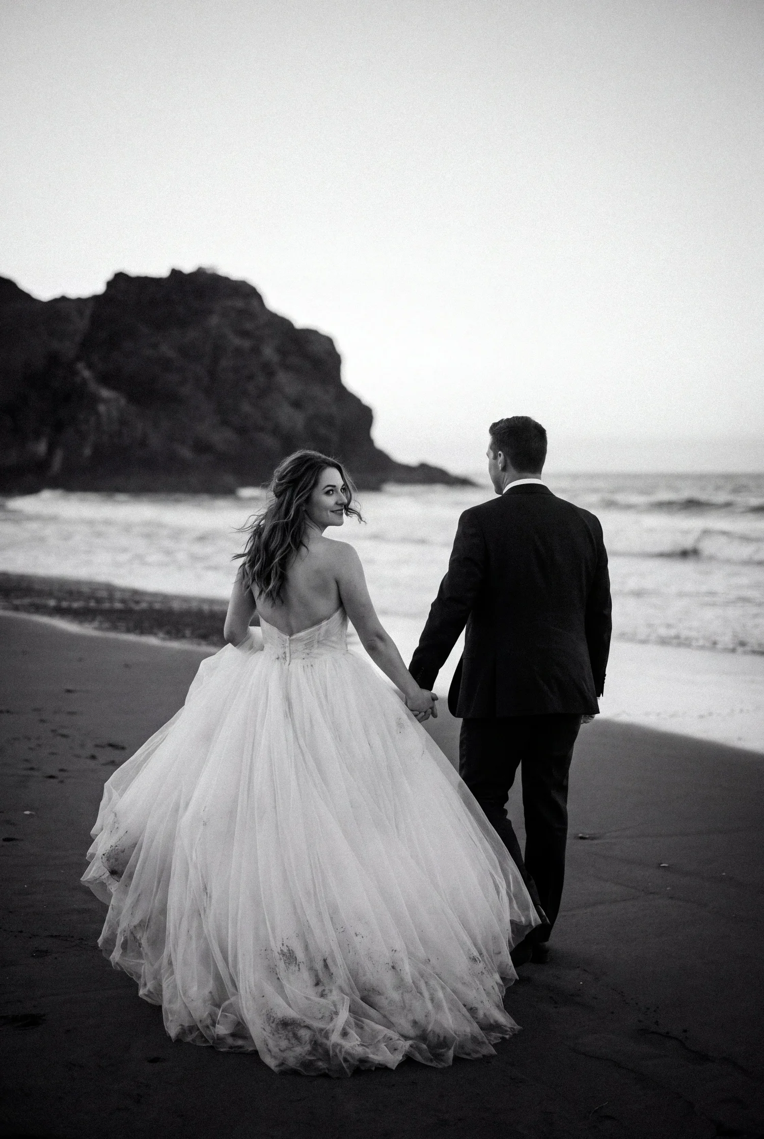 A bride glances back over her shoulder as she and her groom walk hand in hand along a dark sand beach