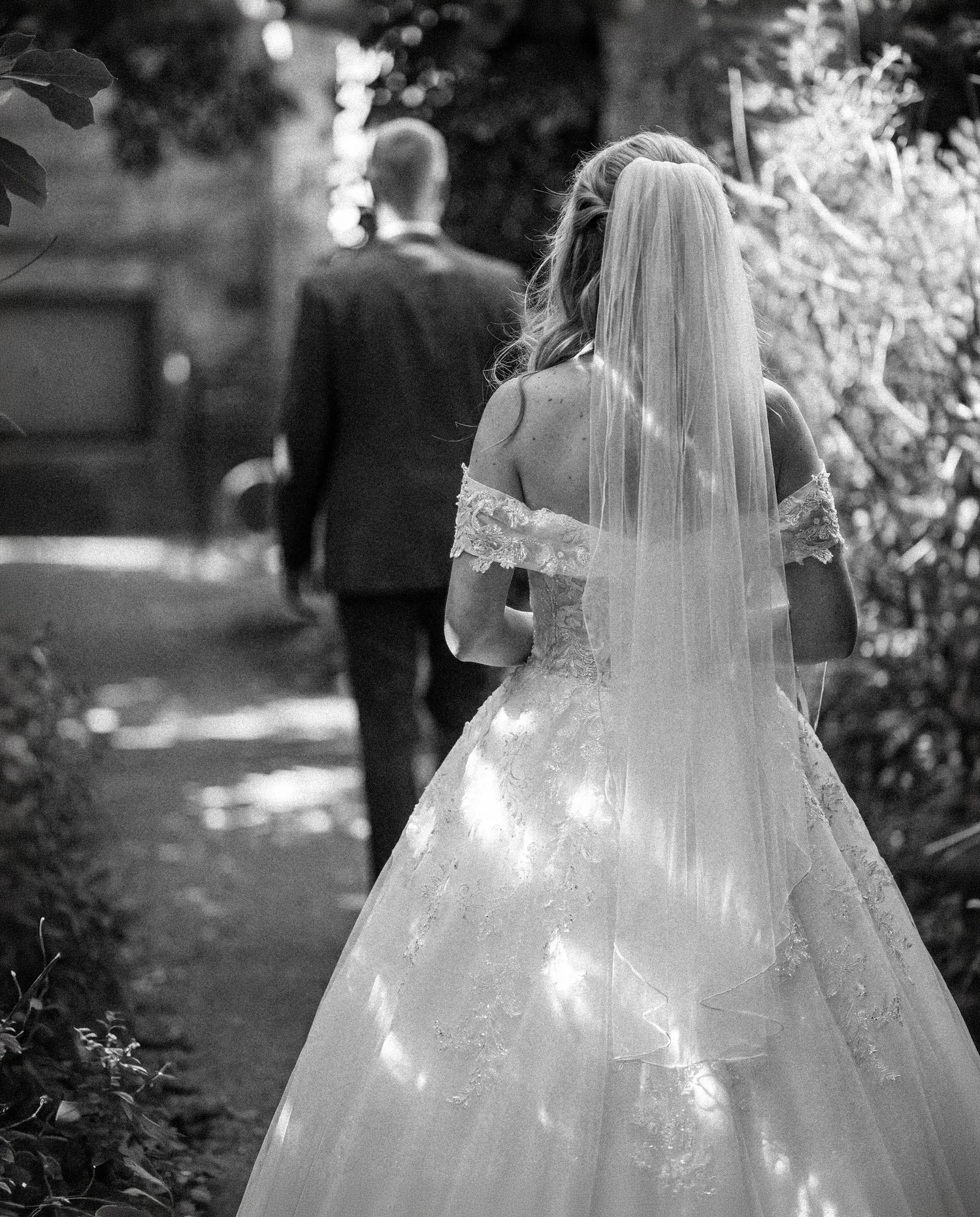 A bride in a lace gown and long veil follows her groom down a shaded garden path