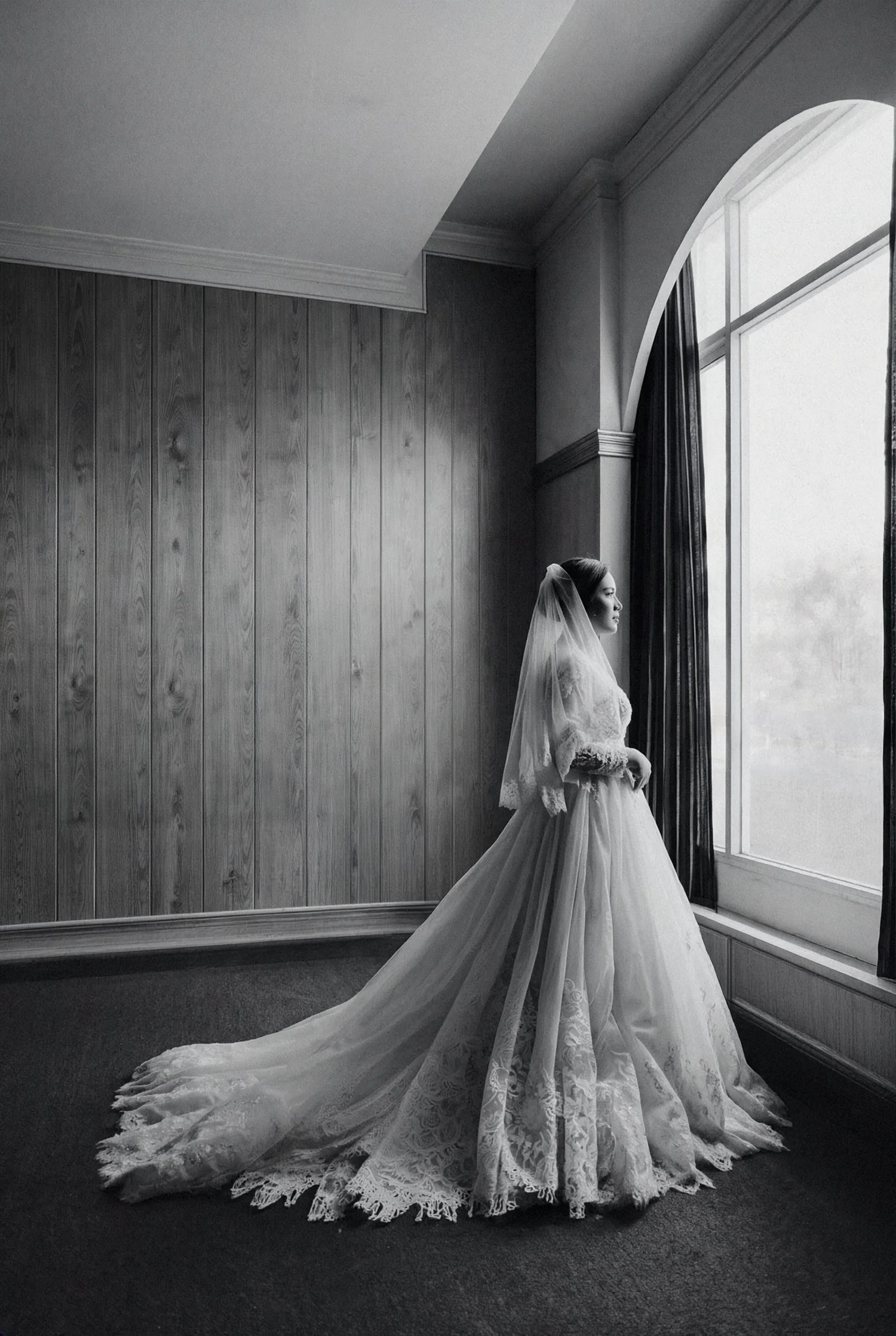 A bride stands alone at a tall arched window, her lace train spread across the floor