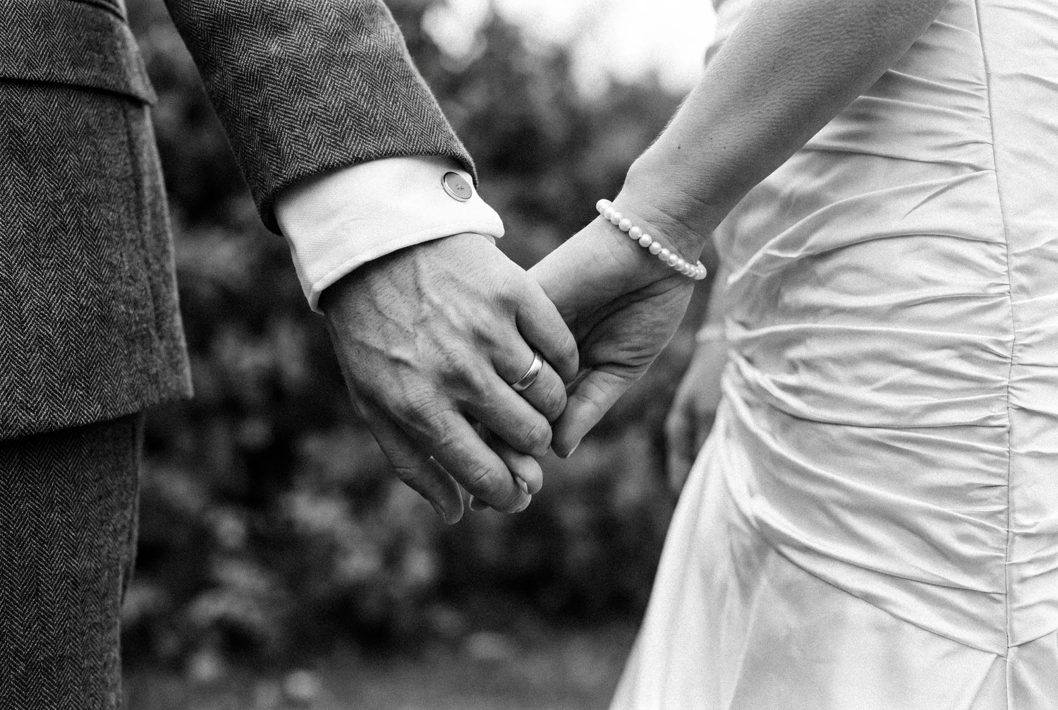 Close detail of a couple holding hands, his wedding band and cufflink beside her pearl bracelet