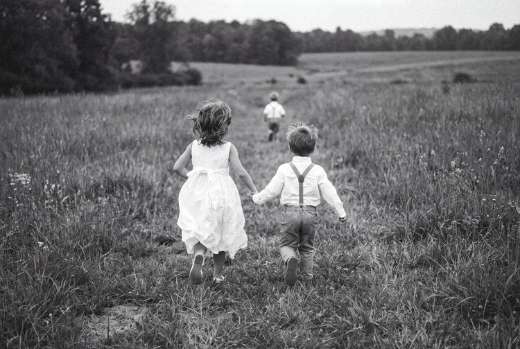 Two young children in wedding clothes run hand in hand across a grassy field