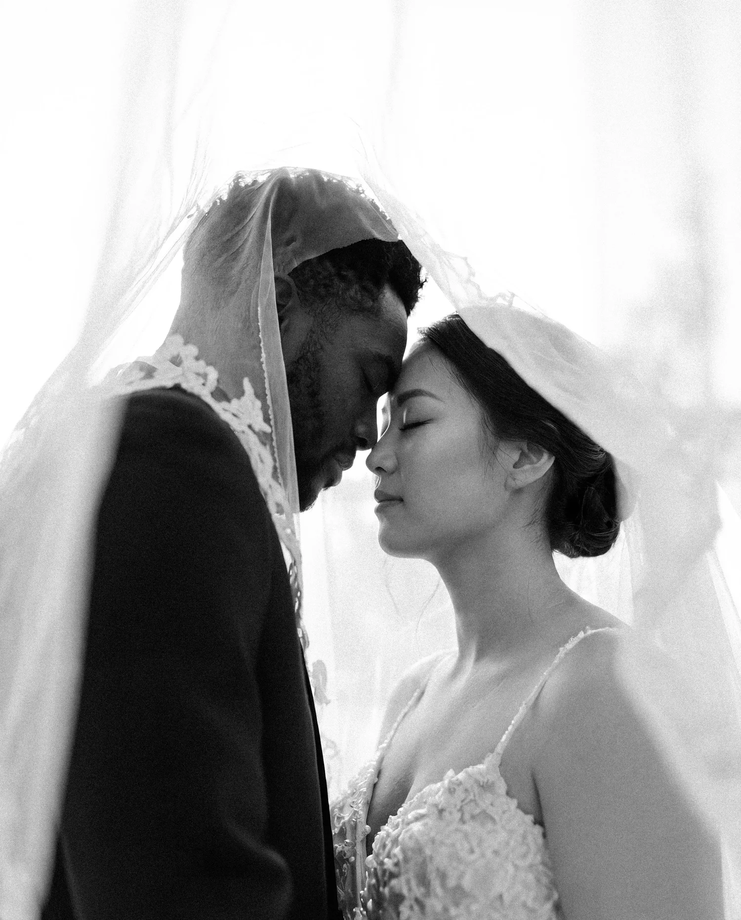 A bride and groom rest their foreheads together beneath her lace veil, eyes closed
