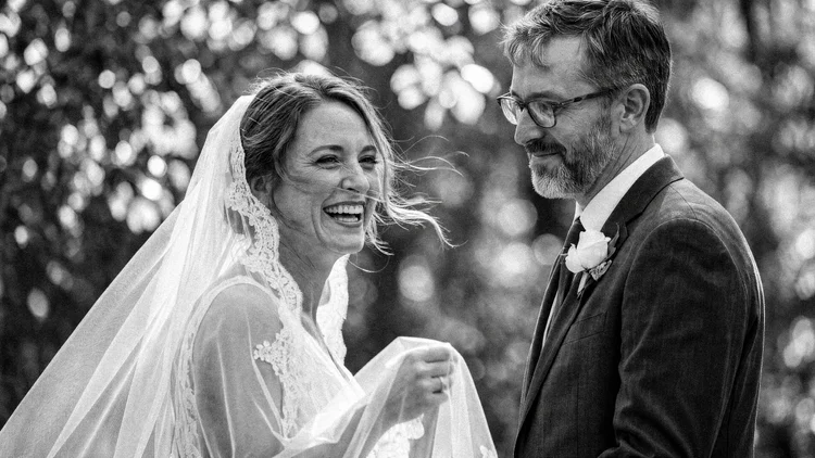 A bride laughs beneath her lace trimmed veil while her groom smiles down at her