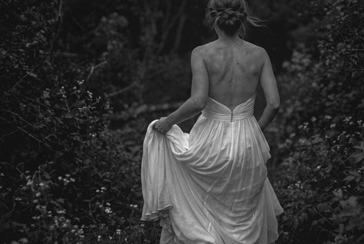 A bride seen from behind gathers her gown as she walks into a dark, overgrown garden
