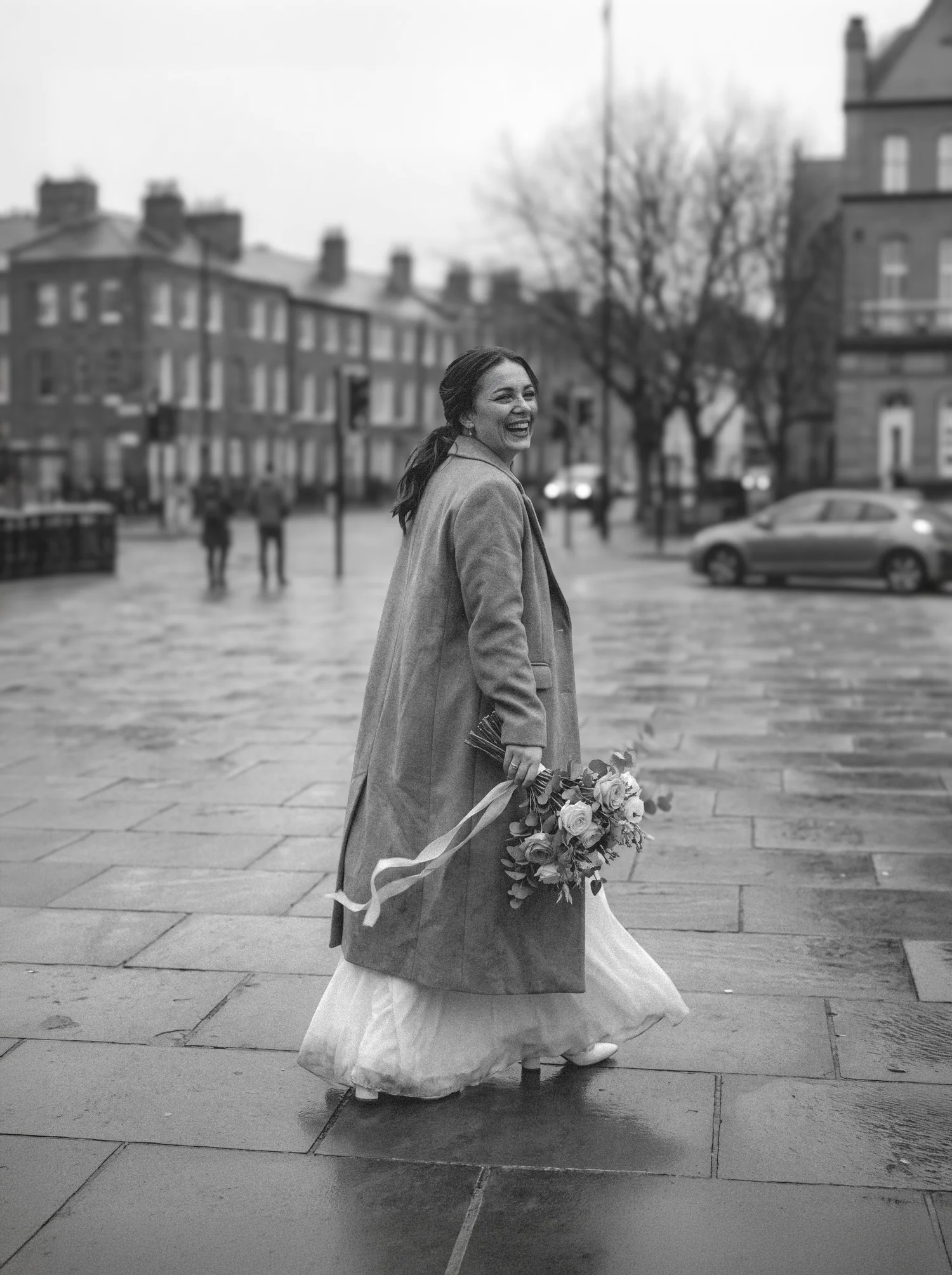 A laughing bride in a long coat over her gown looks back on a wet city street, bouquet in hand