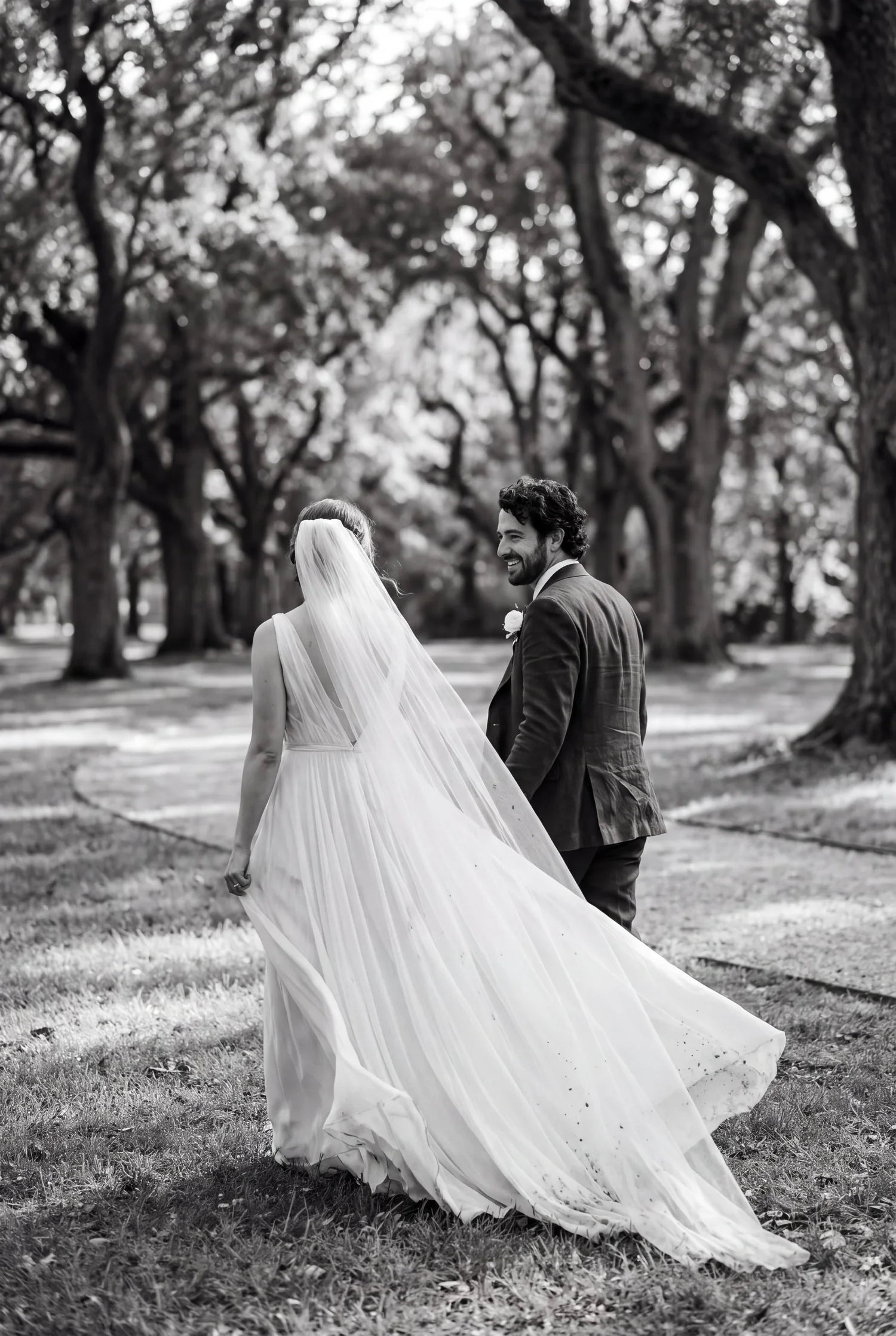 A smiling groom walks beside his bride beneath old oak trees, her long veil drifting behind