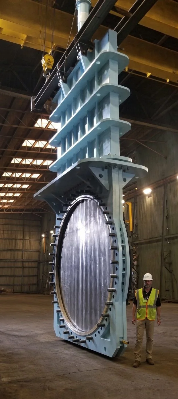 Large fabricated knife gate valve suspended from a shop crane with a worker standing beside it for scale