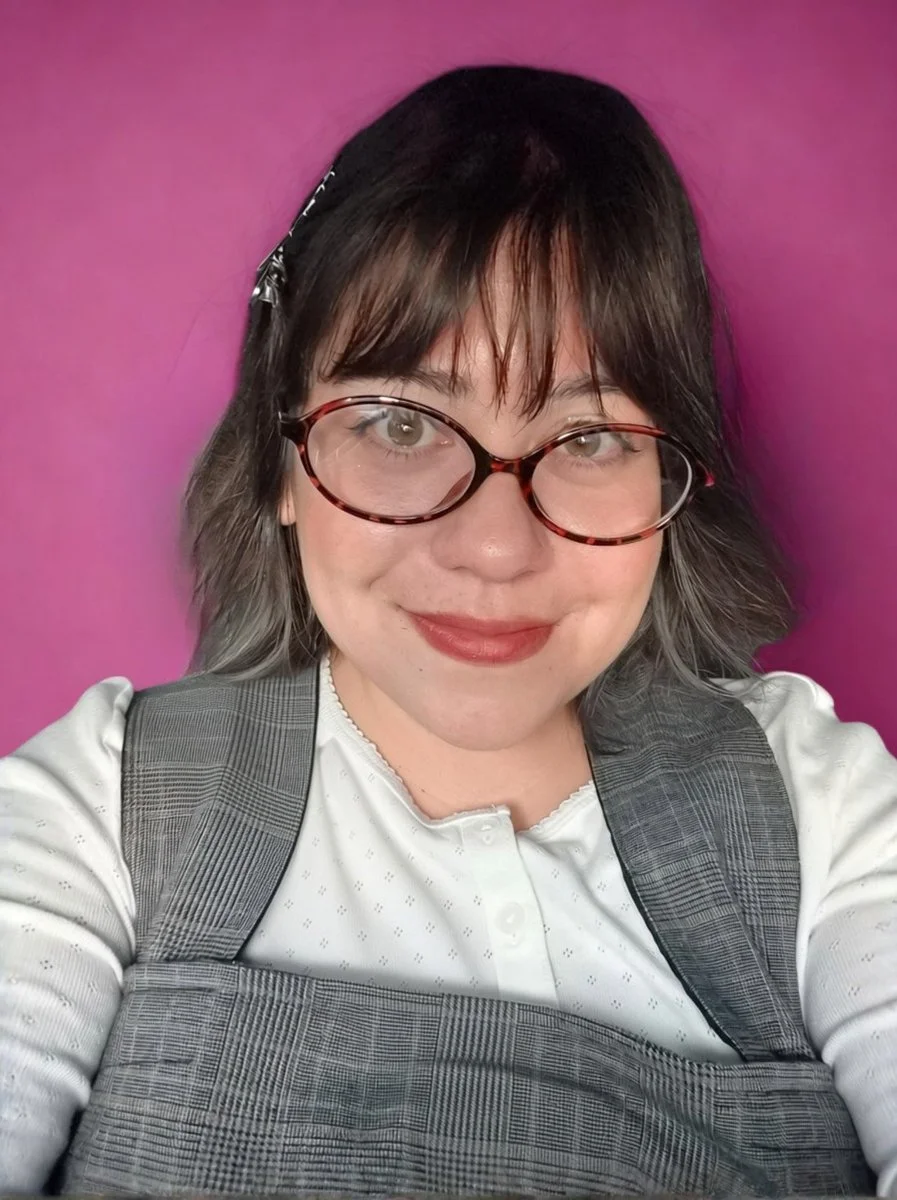 Jess smiling in red oval glasses, a white blouse and a gray plaid vest