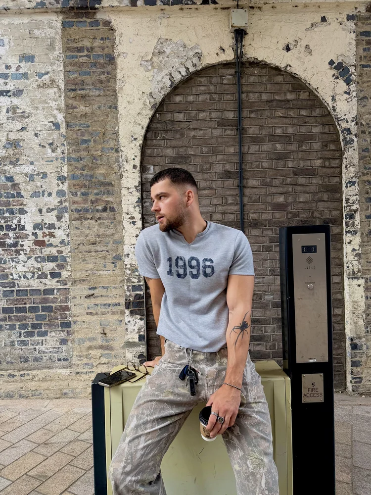 1996 tee and camo trousers against a brick arch