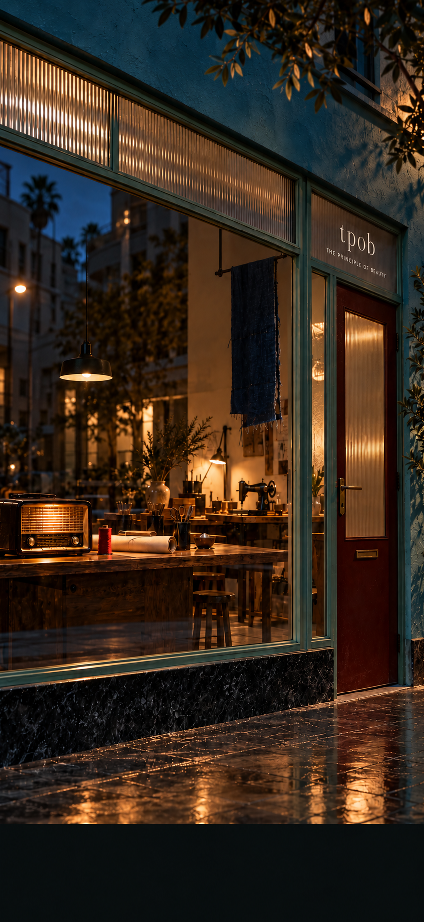 An amber-lit creative studio behind a tall teal storefront, with an antique radio, fabrics, a sewing machine, and a ribbed-glass doorway beneath the TPOB sign.